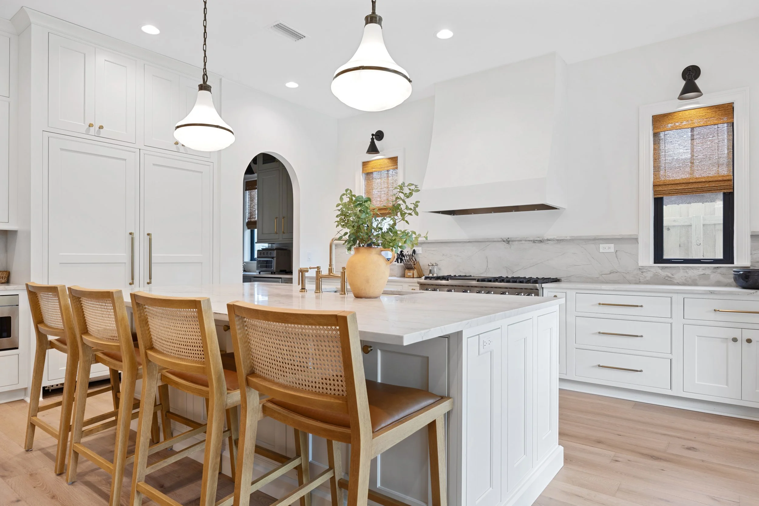 Modern white kitchen with island, wooden bar stools, pendant lights, marble backsplash, and two small windows with bamboo shades.