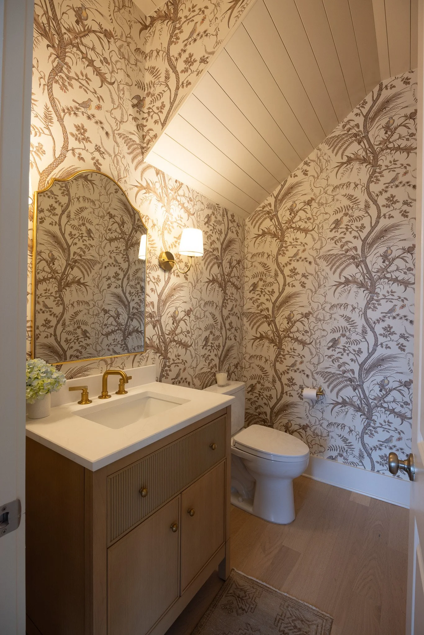 A small bathroom with beige wallpaper featuring tree and bird patterns, a wooden vanity with a white countertop and gold-colored fixtures, a mirror with a gold frame, a wall-mounted light, a toilet, a white plant on the vanity, and a small rug on the wooden floor.