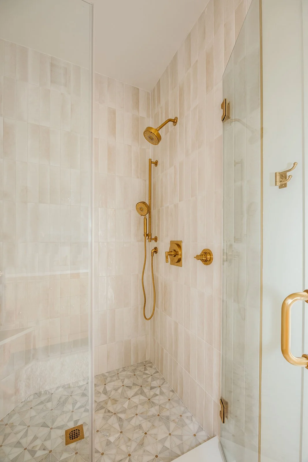 A walk-in shower with gold fixtures, including a showerhead, handheld shower, and control knobs. The shower has beige tiles on the walls and geometric patterned tiles on the floor, with a floor drain visible.