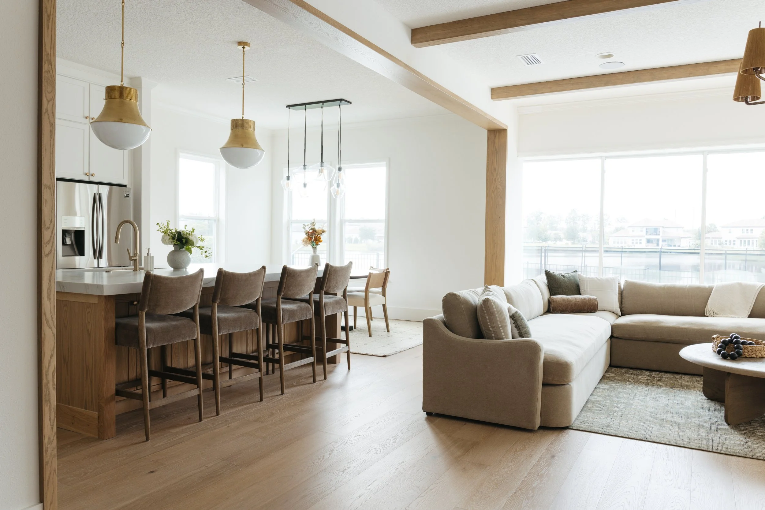 Open-concept living room and kitchen with beige sectional sofa, light wood flooring, white walls, large windows letting in natural light, dining table with chairs, and pendant lights
