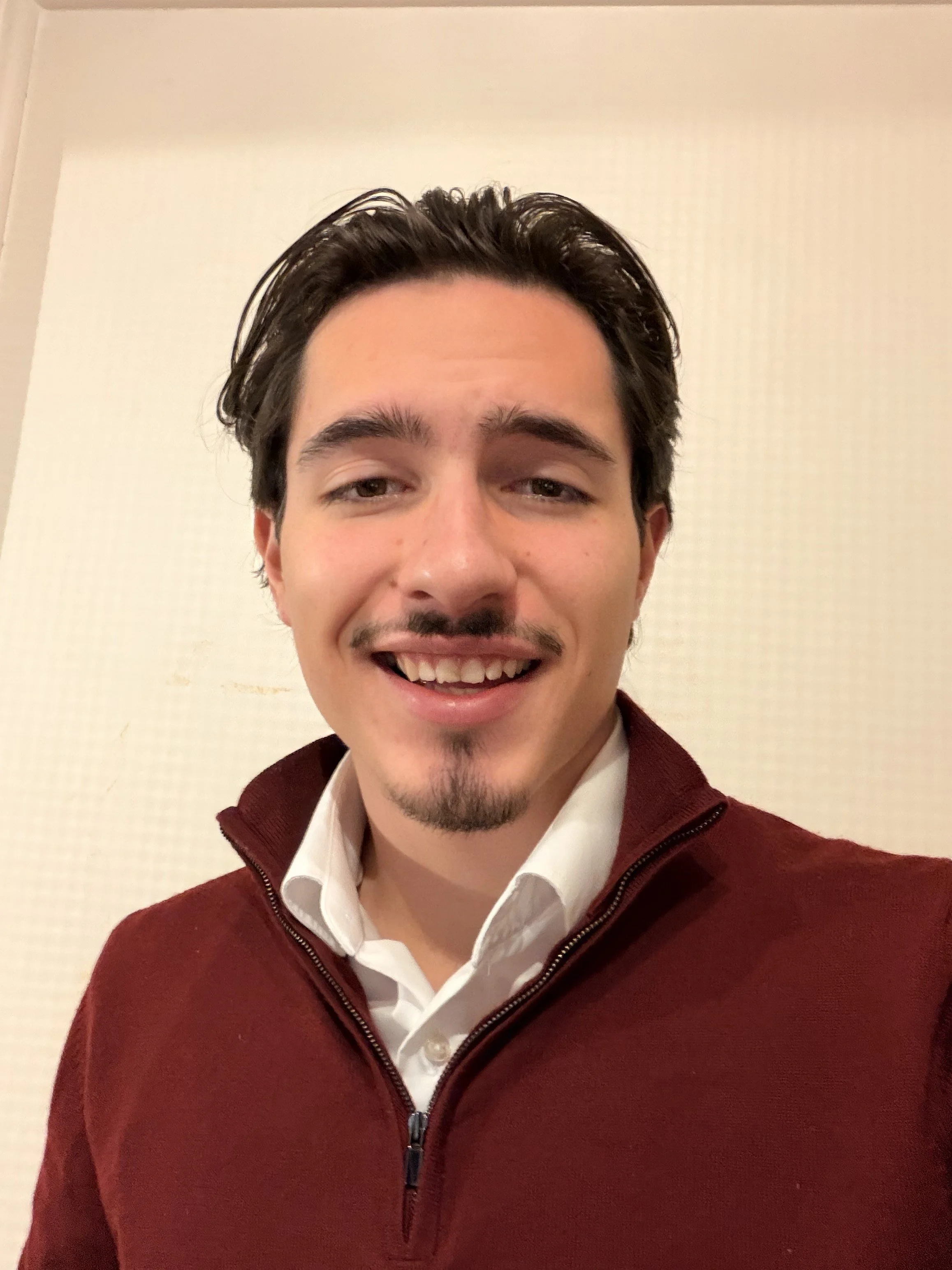 A young man with dark hair, a goatee, and a mustache smiling at the camera, wearing a white collared shirt and a burgundy sweater with a zipper.