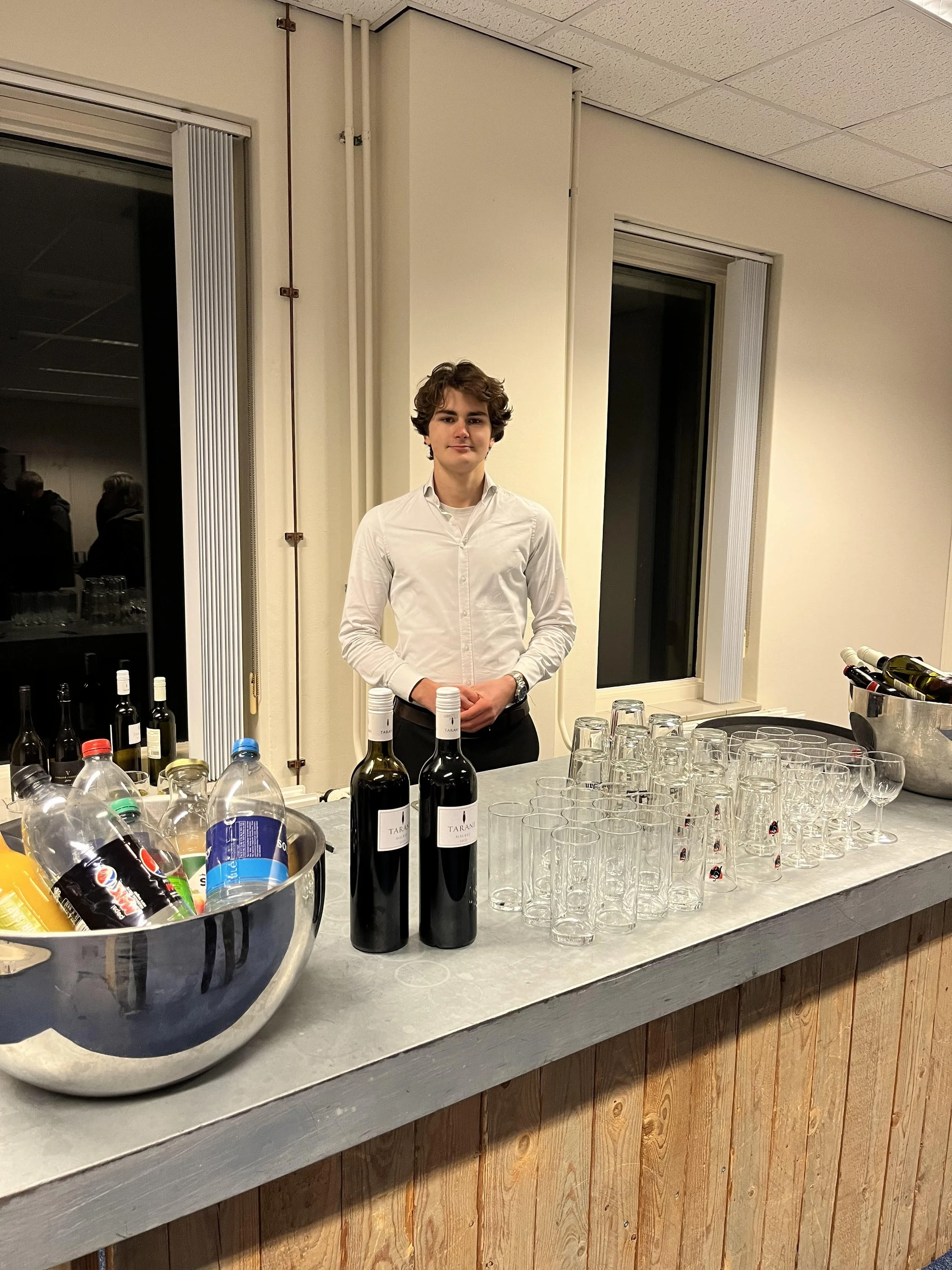 Young man standing behind a bar with wine bottles, glasses, and soft drink bottles on the counter, indoors at night.