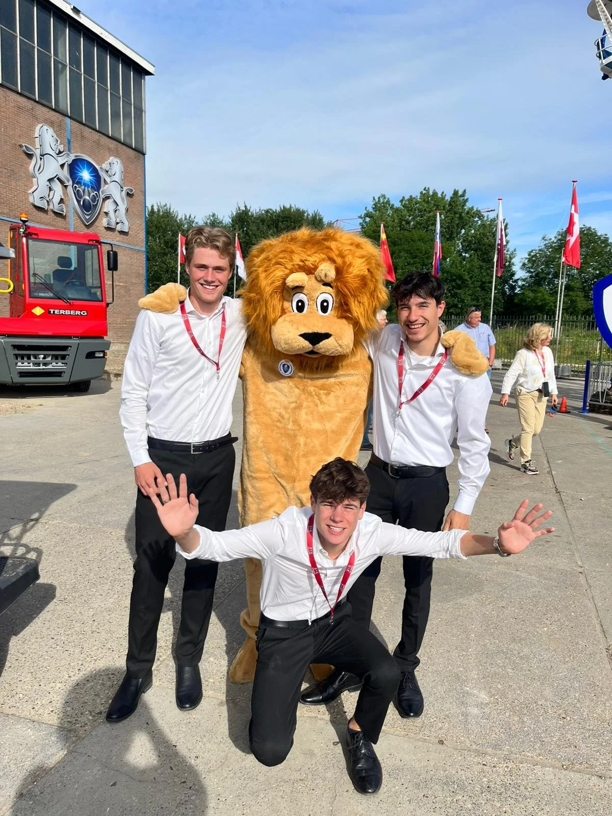 Three young men in white shirts and black pants posing with a person in a lion mascot costume outside near flags and a building with lion emblem. One man kneeling with arms out, two standing with arms around the mascot.