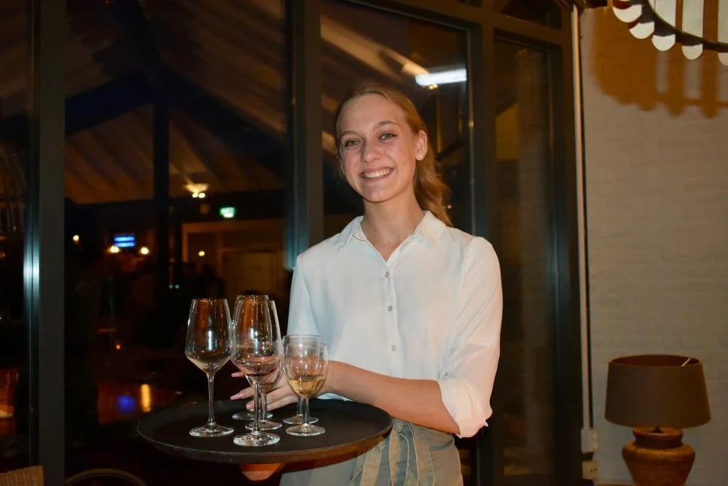 A smiling young woman with red hair holding a tray with four wine glasses at a restaurant or bar.