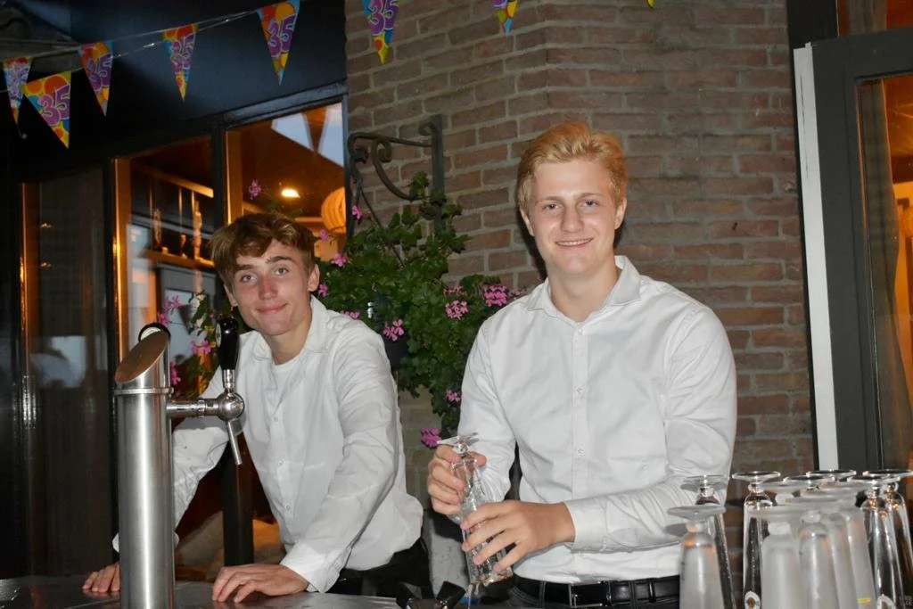 Two young men in white shirts standing behind a bar, smiling at the camera, with colorful party banners overhead and flowers on the wall behind them.
