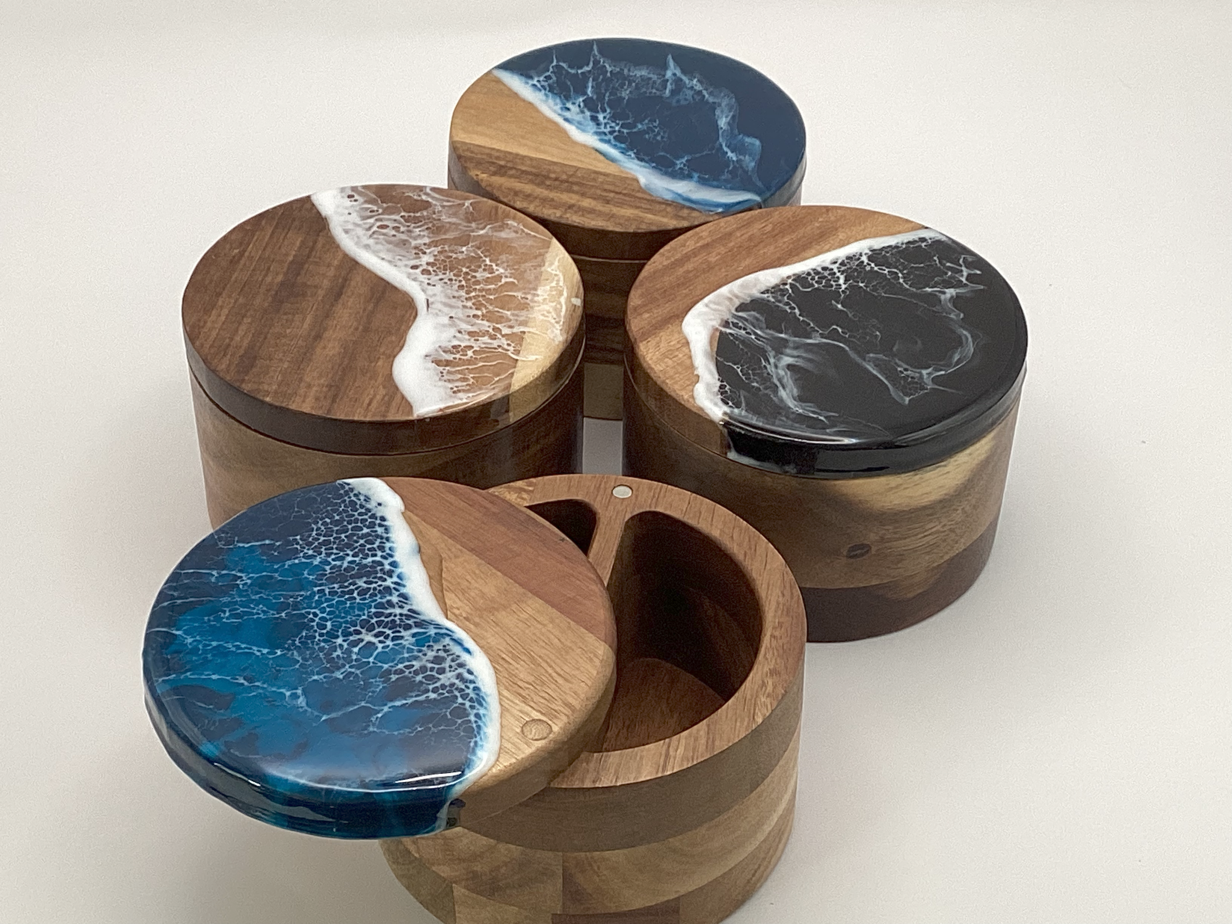 Set of five round wooden boxes with ocean wave resin-inspired designs on the lids, in shades of blue, black, white, and brown, arranged on a white surface.
