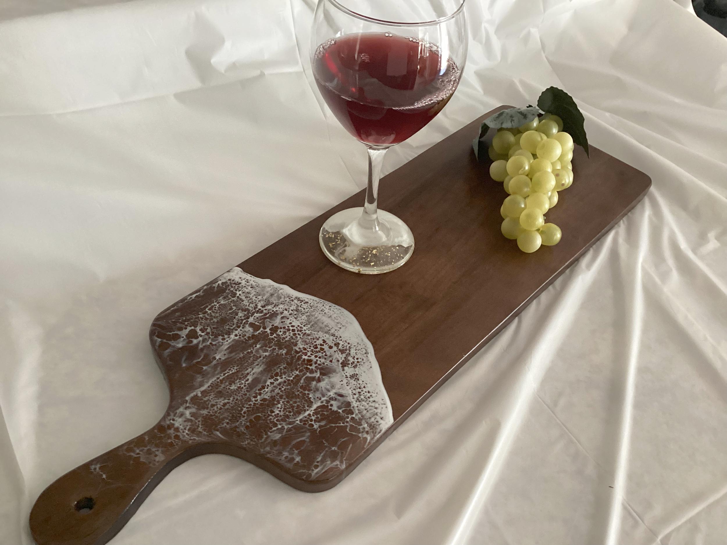 A wooden serving board with a bunch of green grapes, a glass of red wine, and a soap-like decoration resembling a foam of beer placed on a white cloth surface.