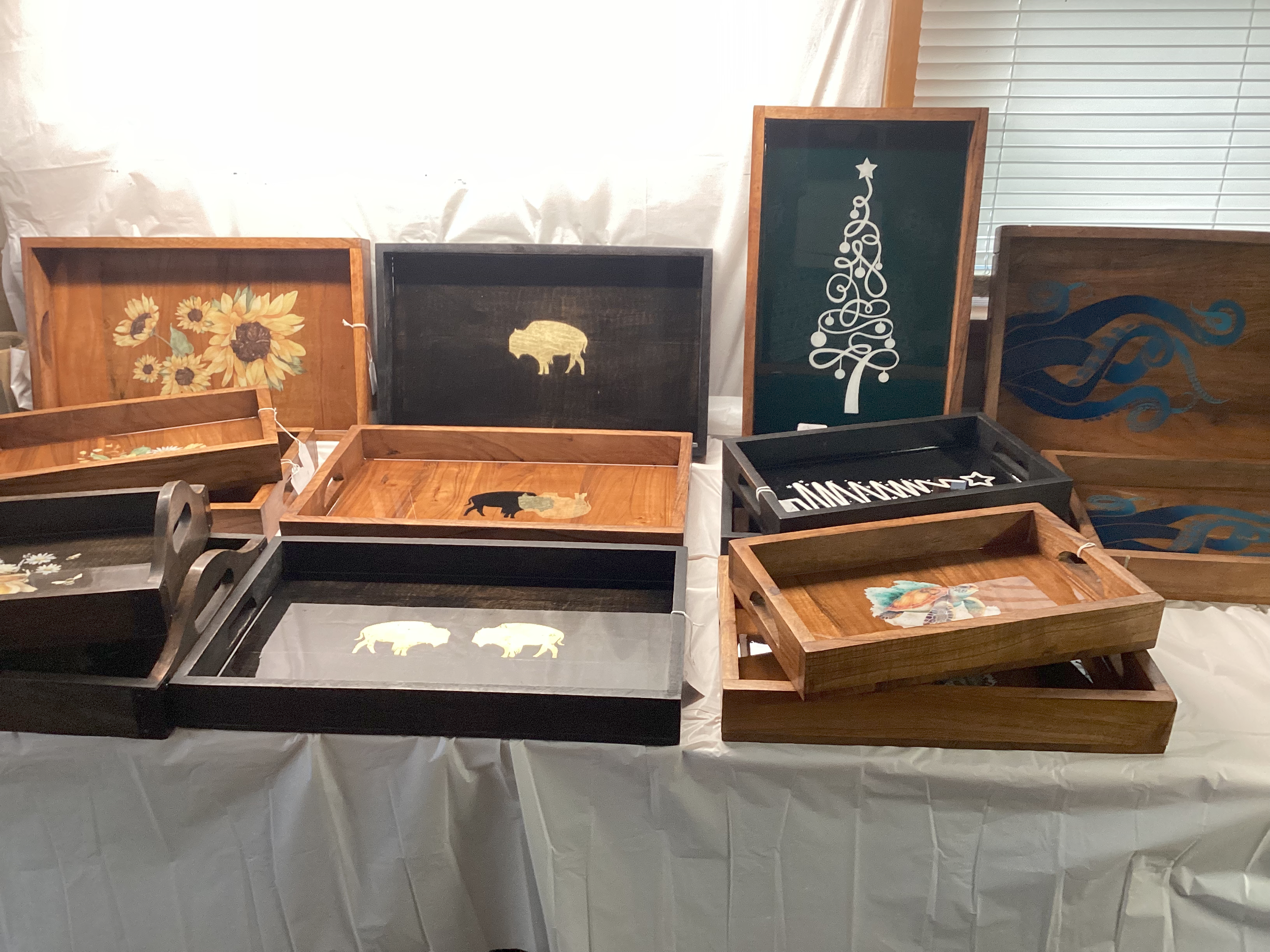 Display of wooden trays with painted designs, including sunflowers, a buffalo, and abstract blue patterns, arranged on a white tablecloth at craft fair.