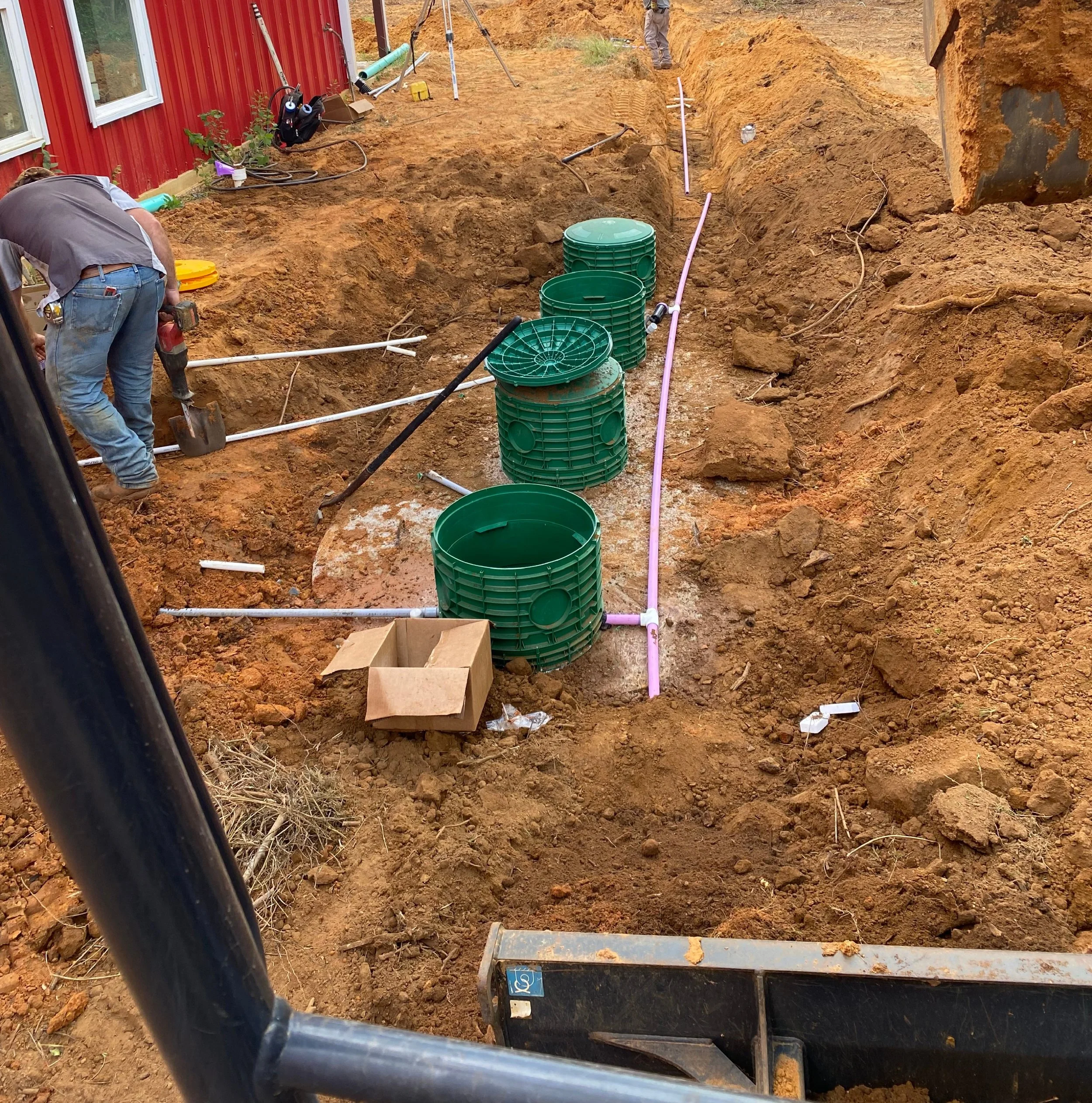 Construction site with green septic tank risers, pink piping, and workers installing underground plumbing next to a red building.