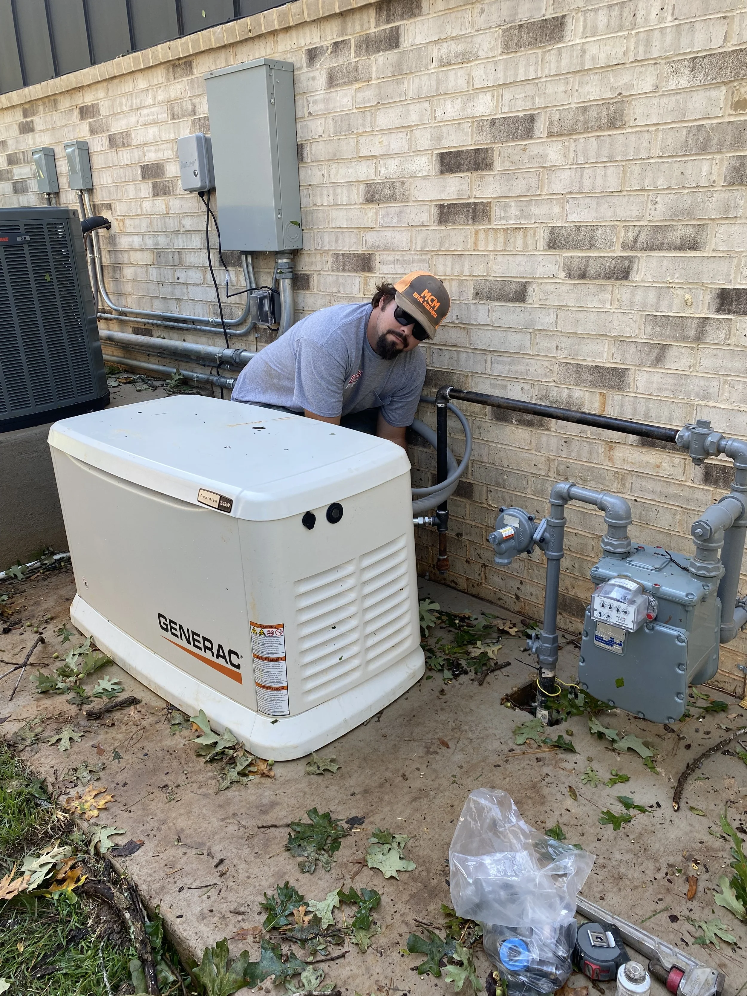 A man wearing sunglasses, a gray t-shirt, and an orange and gray cap crouches next to a generator outside a building. The area has various pipes, electrical panels, and tools on the ground surrounded by some fallen leaves.