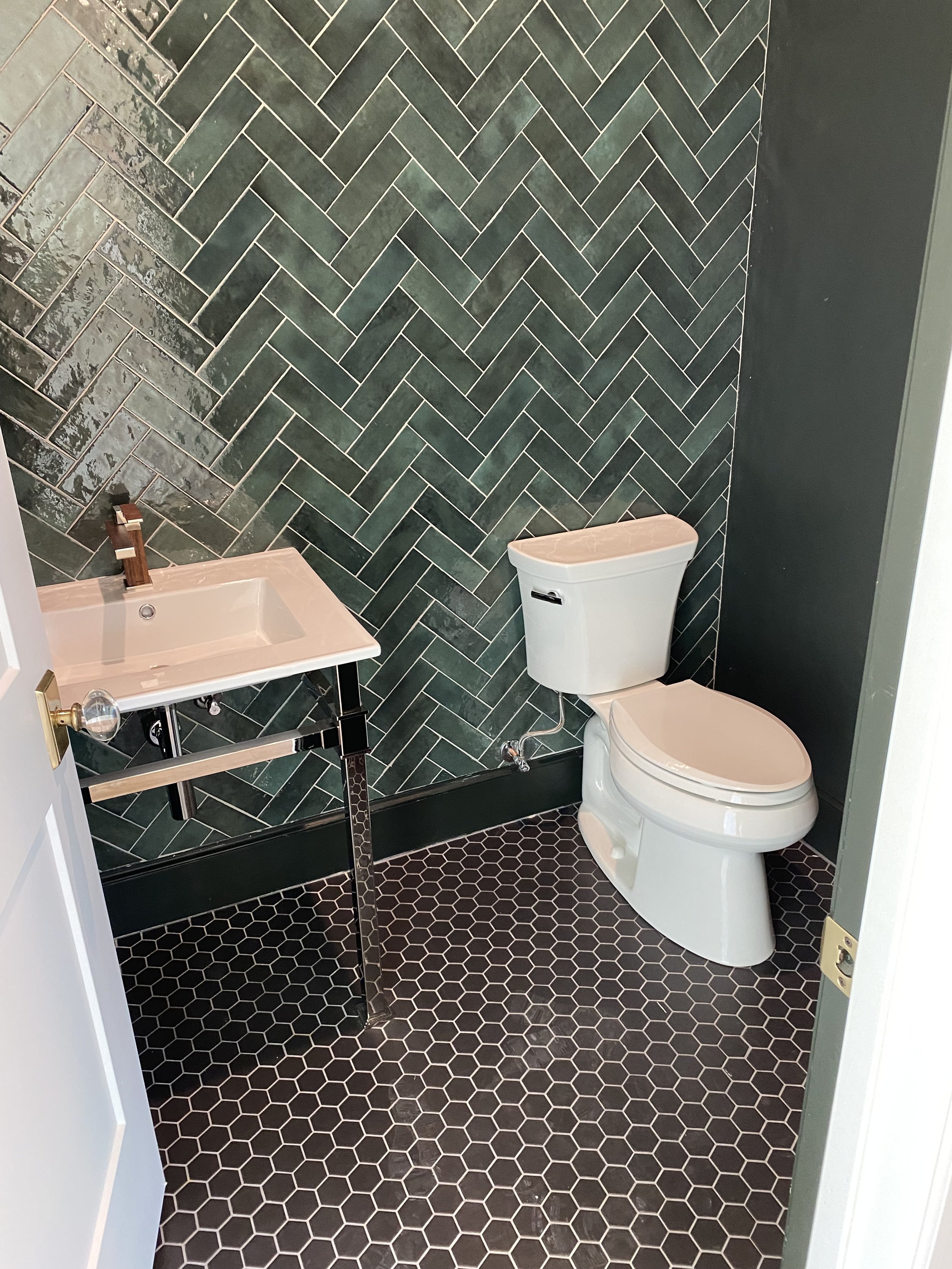 Small bathroom with black hexagon floor tiles, dark green herringbone wall tiles, and a white toilet and sink.