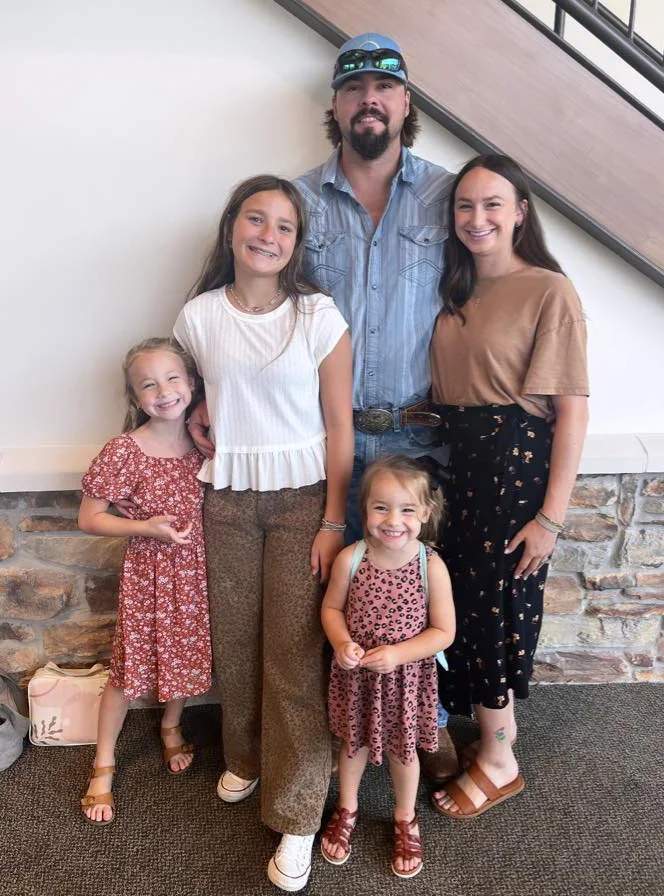 Meet the Minks! A family of five, two adults and three young girls, standing together smiling inside a building with a stone wall and staircase in the background.