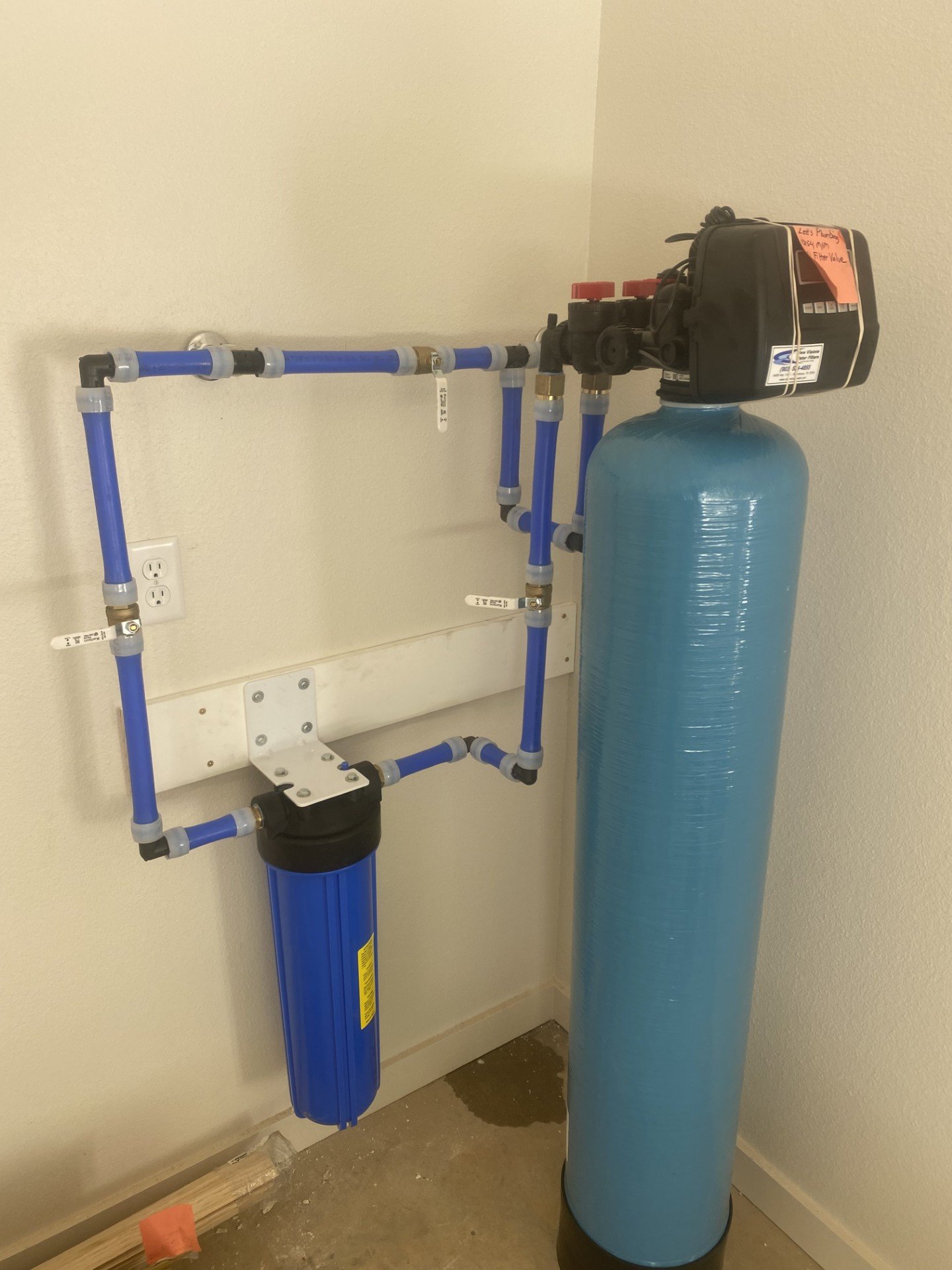 Water filtration system with blue tank, piping, and control valve mounted on a wall.