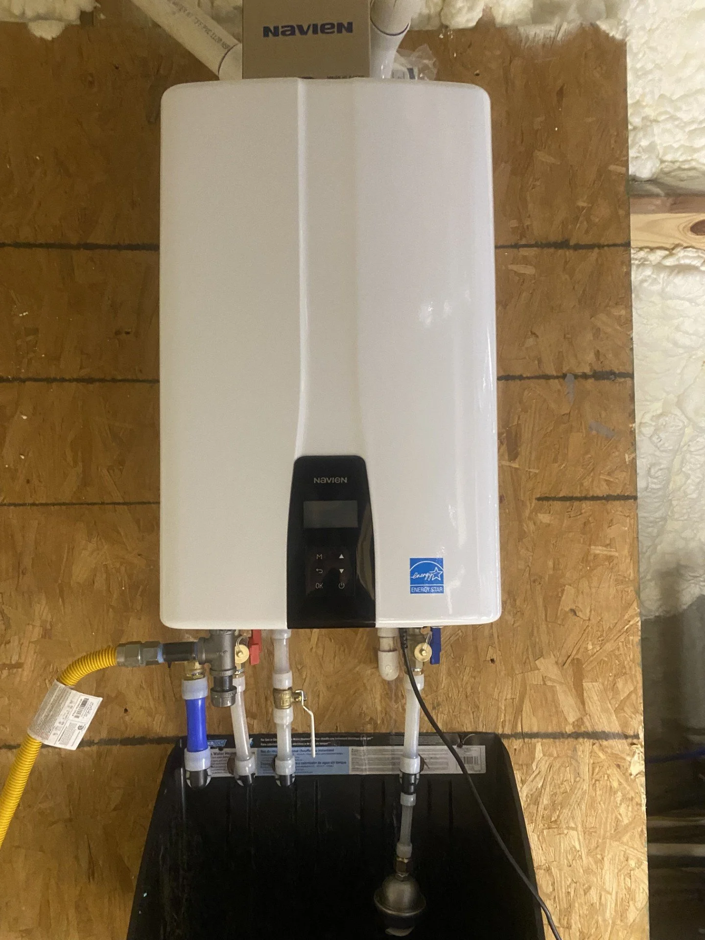 An electric tankless water heater mounted on a wooden wall with various pipes and hoses connected to it.