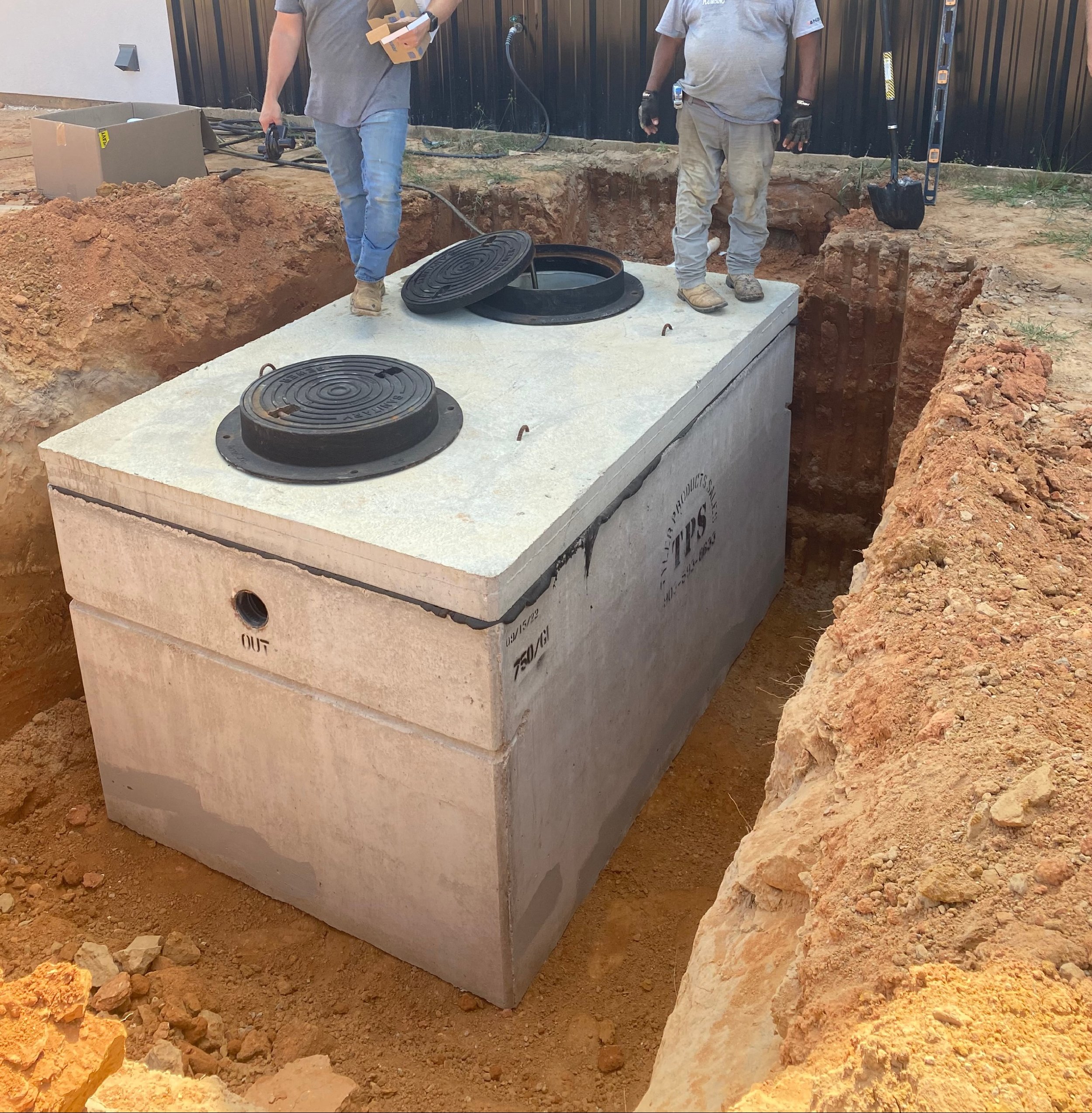 Construction workers installing a large underground storage tank, with open manhole covers, in a dirt excavation site.