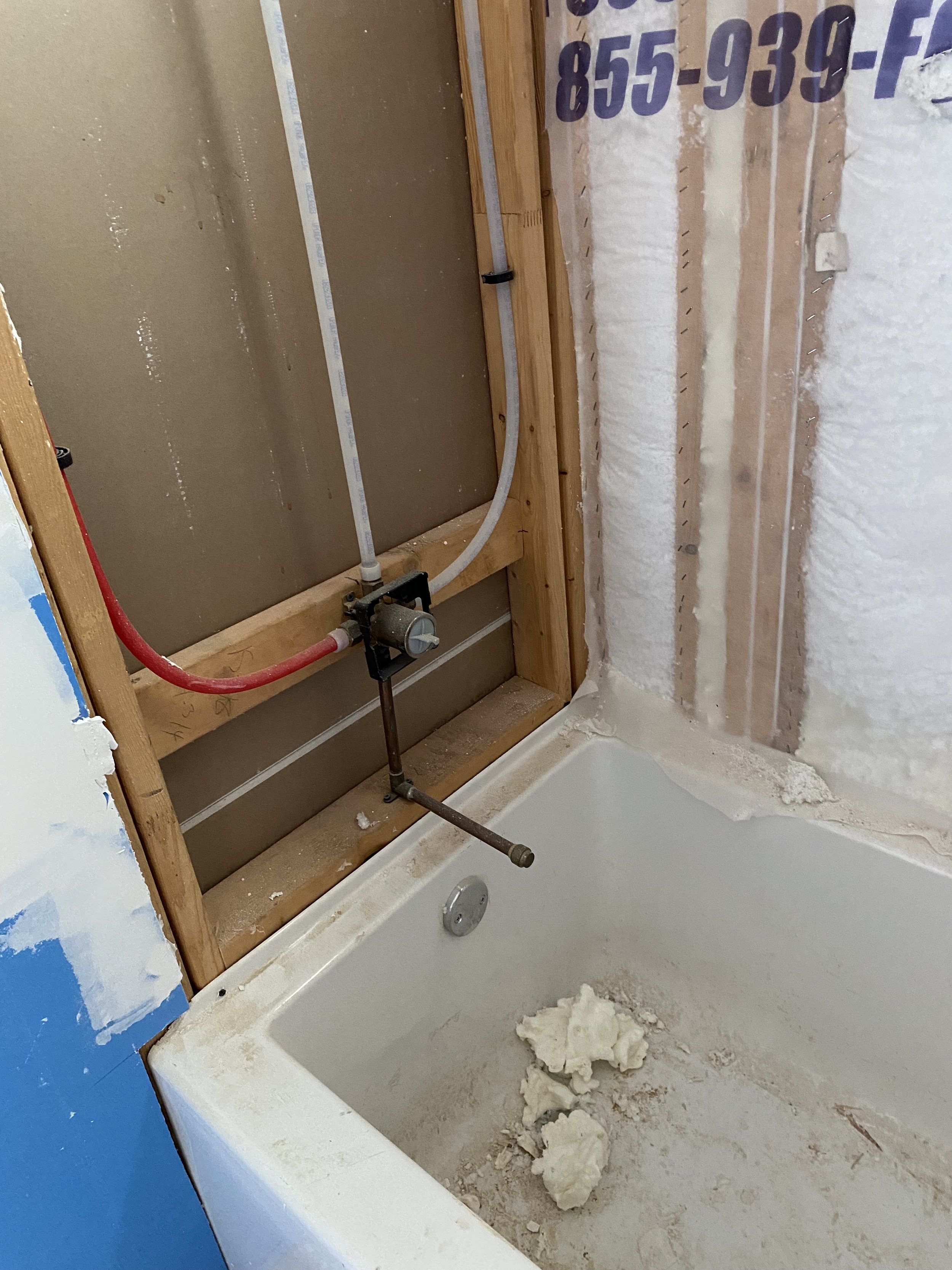 Bathroom under renovation showing an empty bathtub with some debris, exposed plumbing, and partial wall framing with insulation and drywall