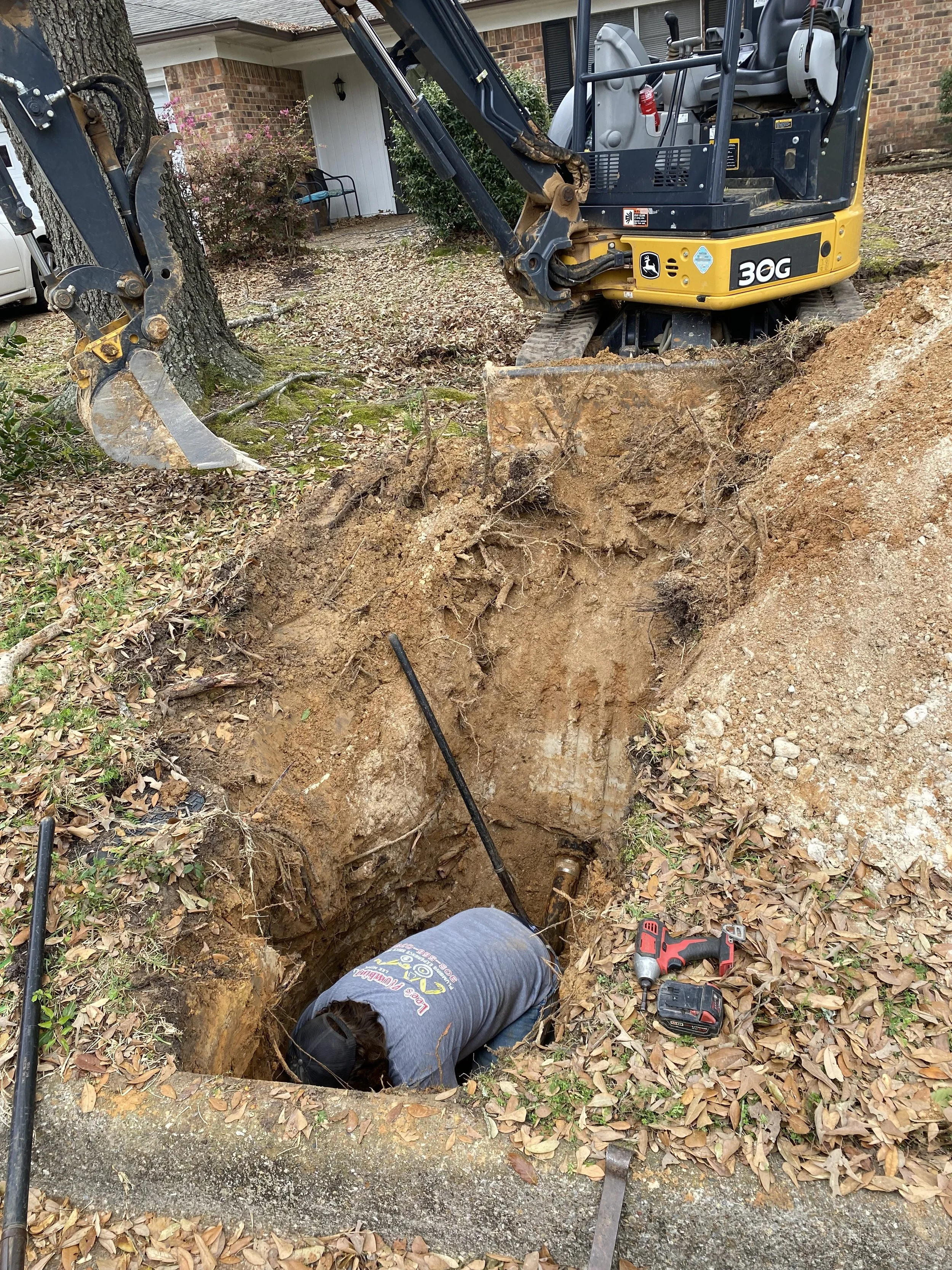 A construction worker is working inside a deep hole in the ground, surrounded by dirt and leaves, with a small yellow and black excavator nearby. The worker is wearing a gray shirt and a black cap, leaning into the hole, with tools like a cordless drill and metal rods around.