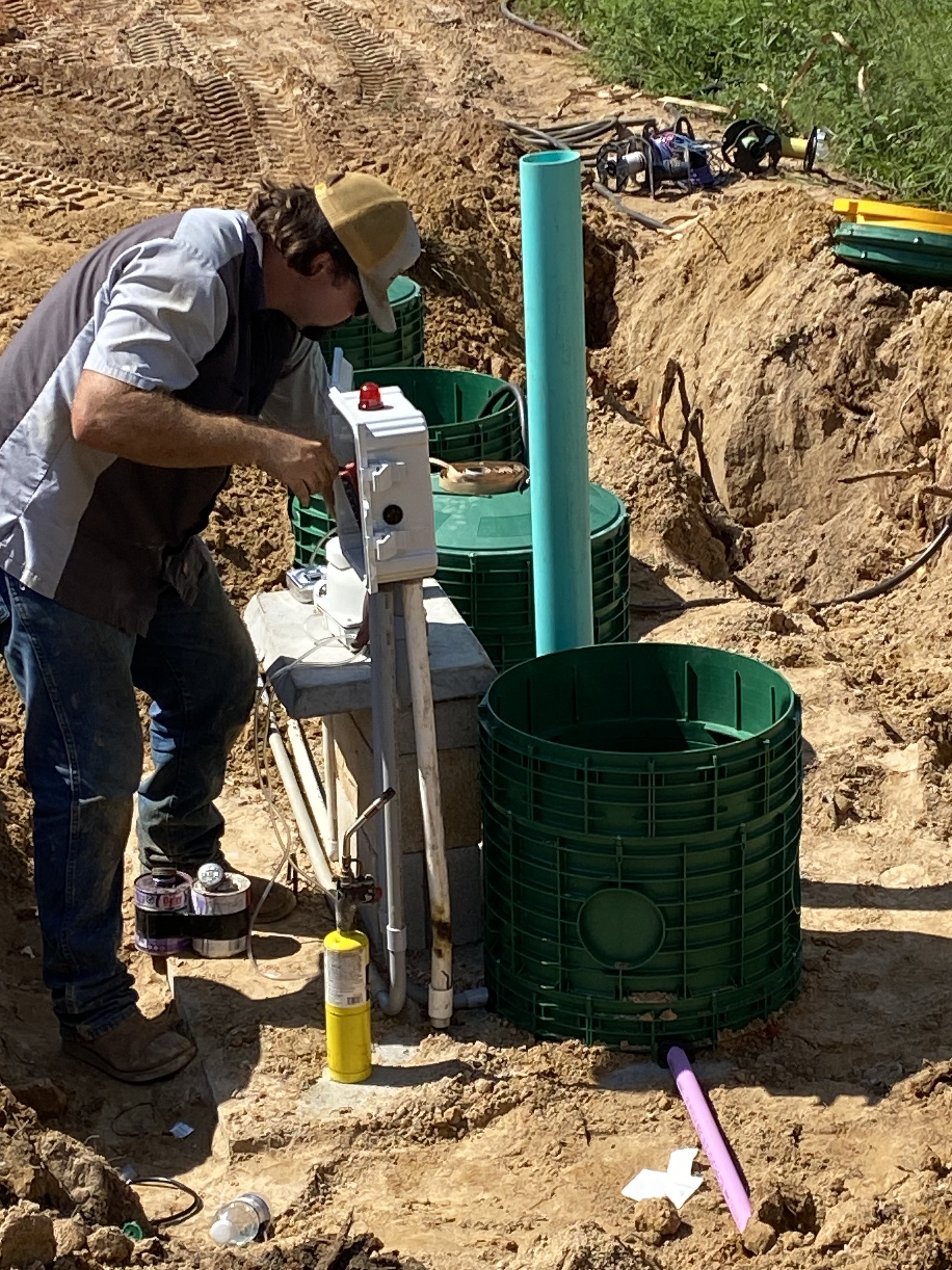 A man working on a well or water system installation in a dirt outdoor area with pipes, tools, and equipment.