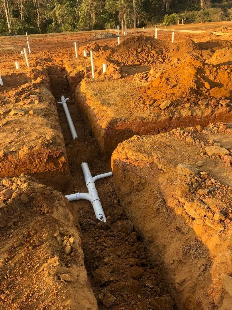 Construction site with dug trenches and white PVC pipes installed in the soil, preparing for underground utility installation.