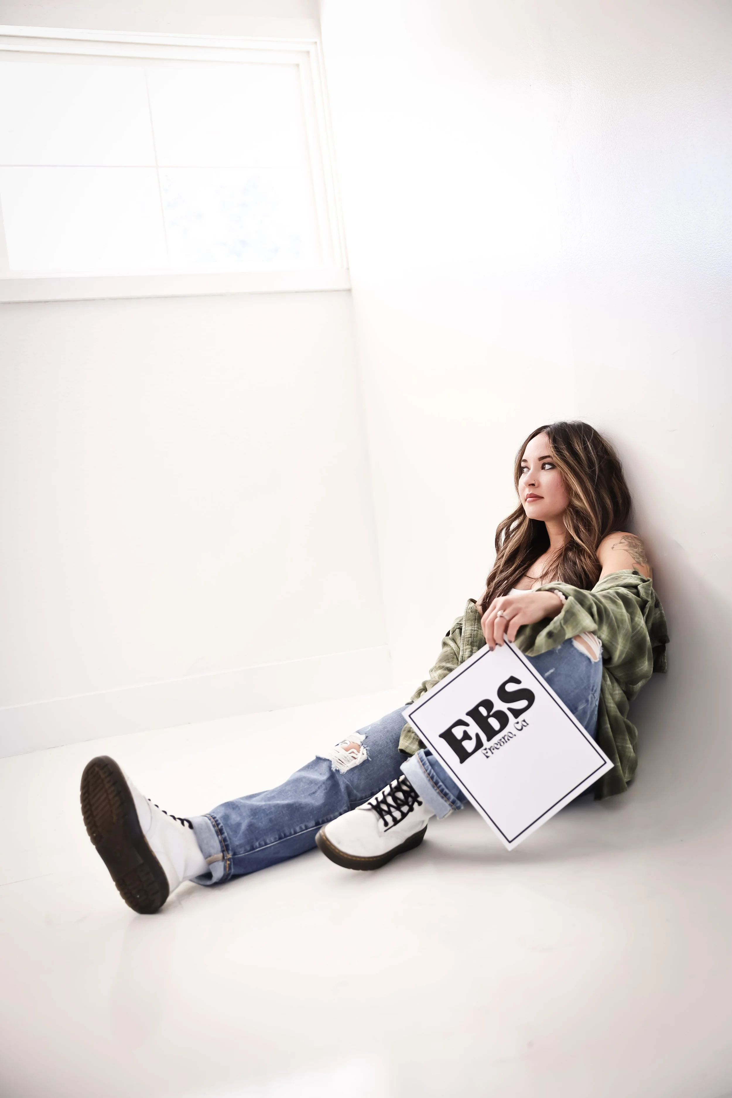 Esthetician seated against a curved white wall holding an EBS Fresno, CA sign, looking off to the side.