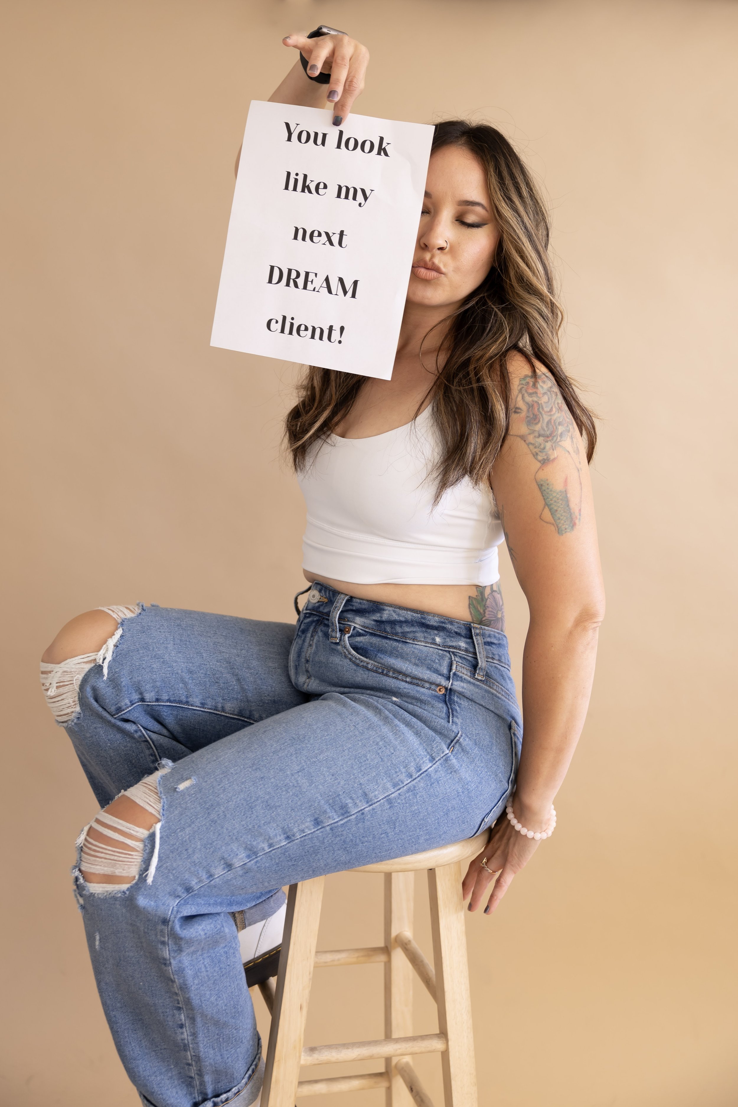 Esthetician posing on a wooden stool and holding a sign that reads “You look like my next DREAM client!”