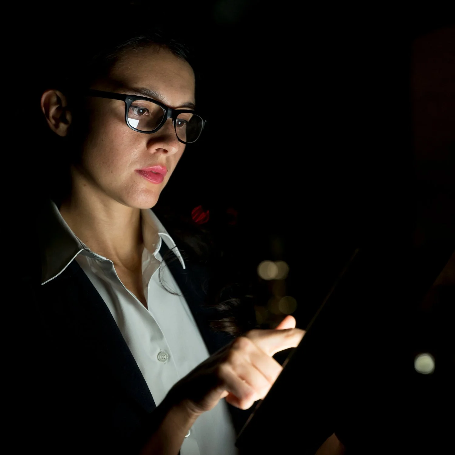 Woman in glasses using a tablet at night with a dark background.