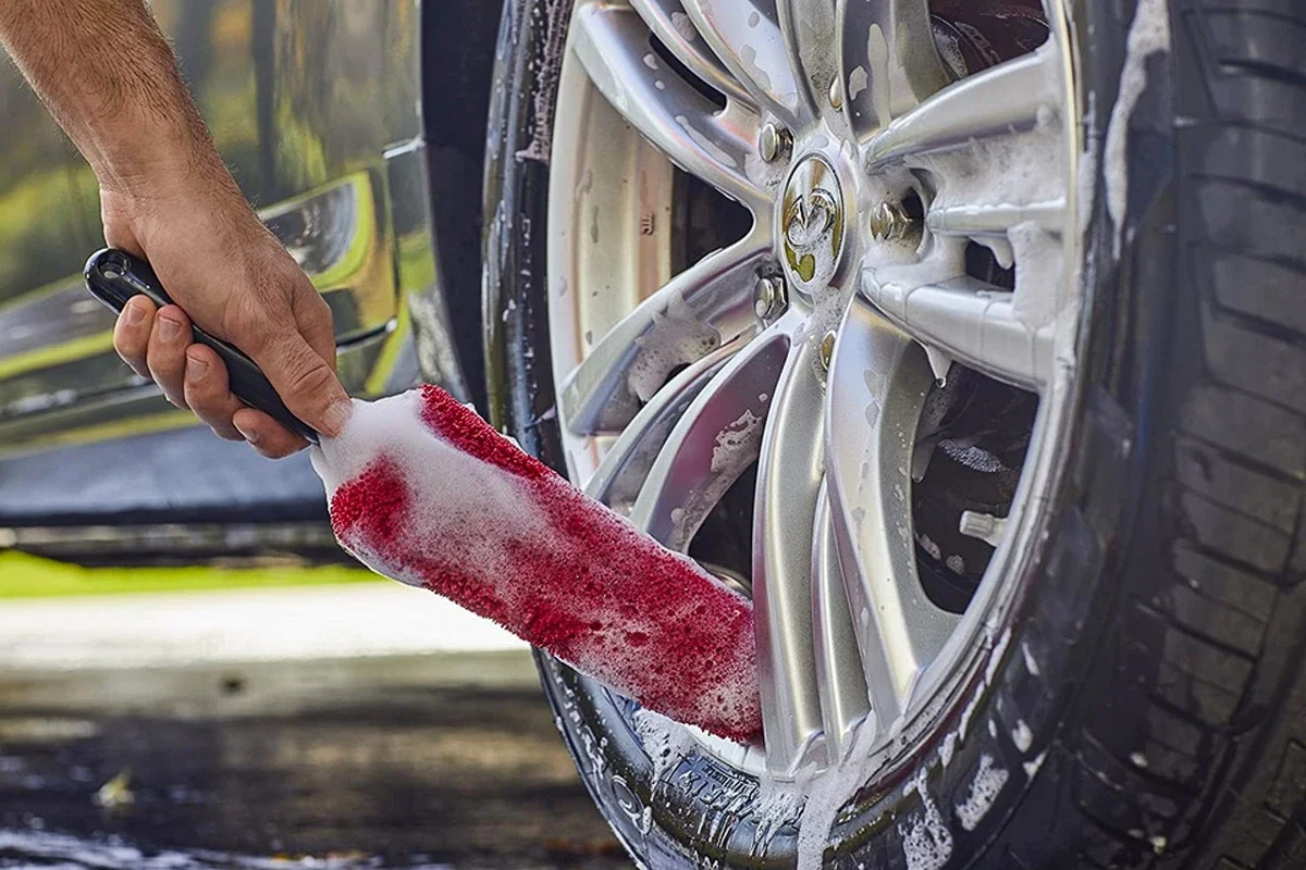 A person washing a car wheel with a red soap sponge at an outdoor car wash station.