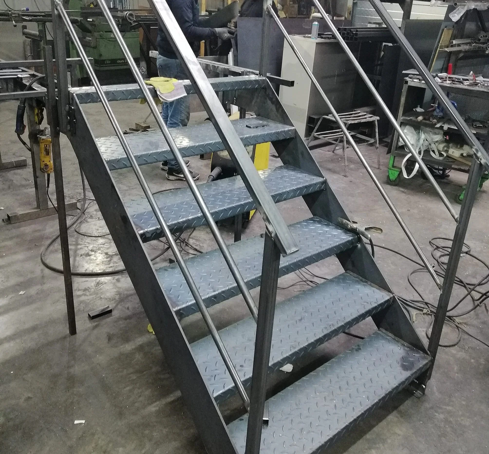 Escalier en métal avec marches en acier à rainures, dans un atelier de fabrication, avec deux hommes travaillant en arrière-plan.