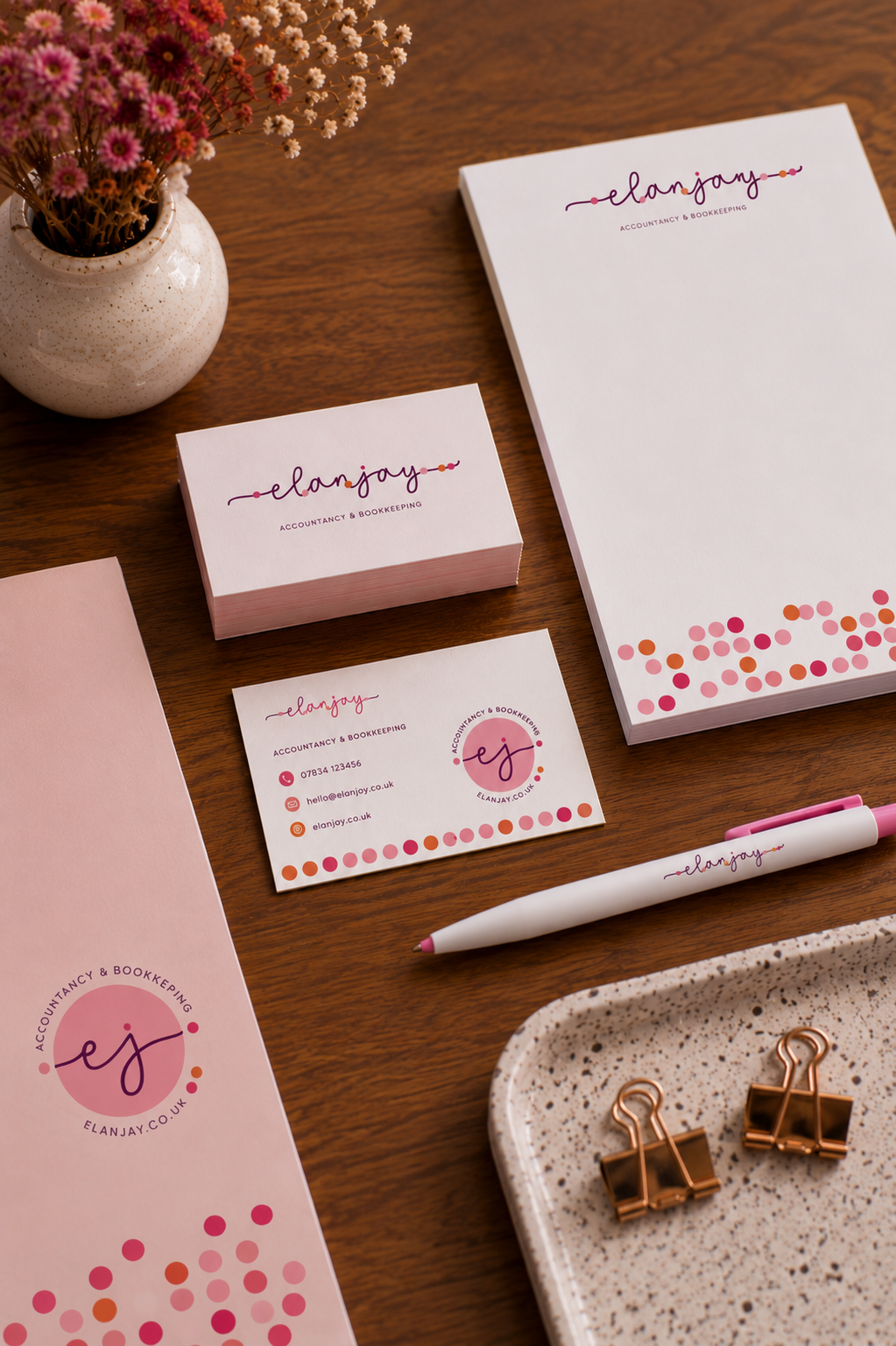 Business branding items for Elanjay Accountancy & Bookkeeping, including a business card, a notepad, a pen, and a box of sticky notes, arranged on a wooden desk with a vase of pink and white flowers.