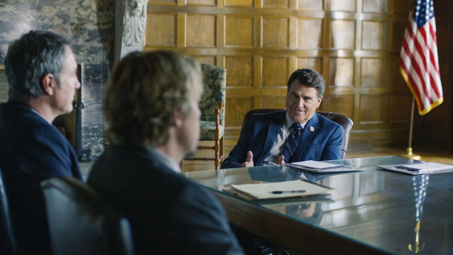 A man in a blue suit and tie sitting at a large desk, engaging in a conversation with two other men. He is smiling and gesturing with his hands. An American flag is visible in the background.