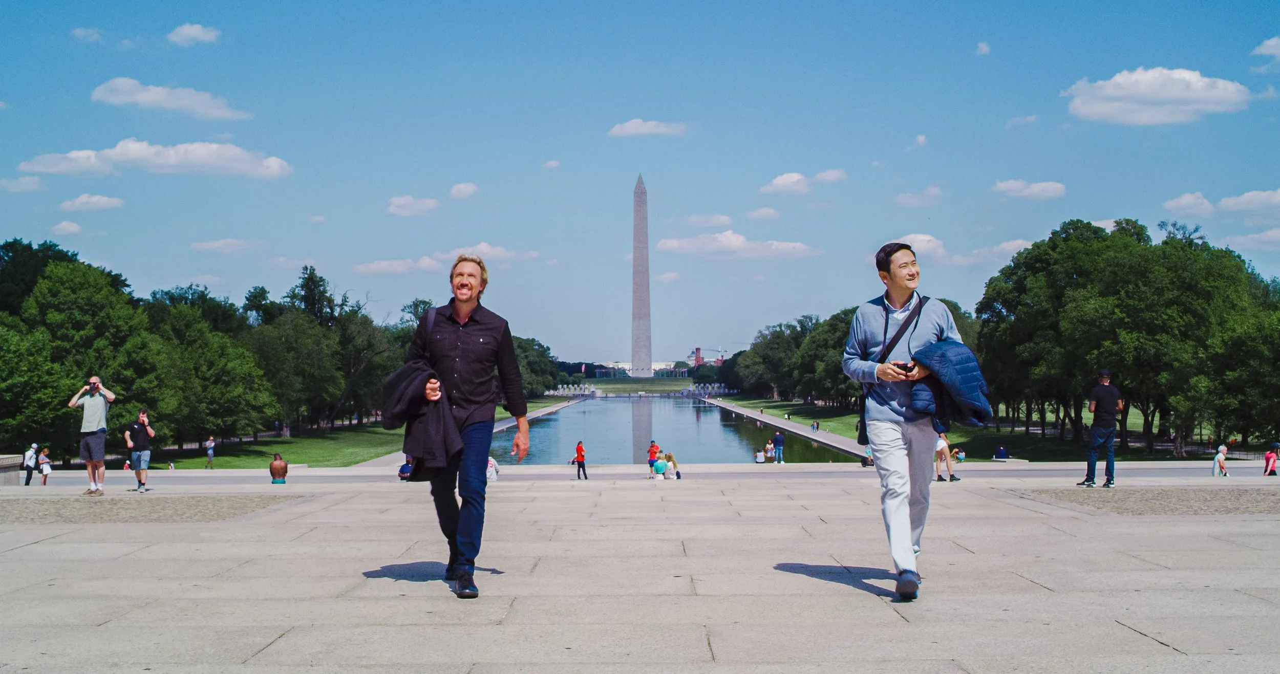 People walking in front of the Lincoln Memorial Reflecting Pool with the Washington Monument in the background on a sunny day.