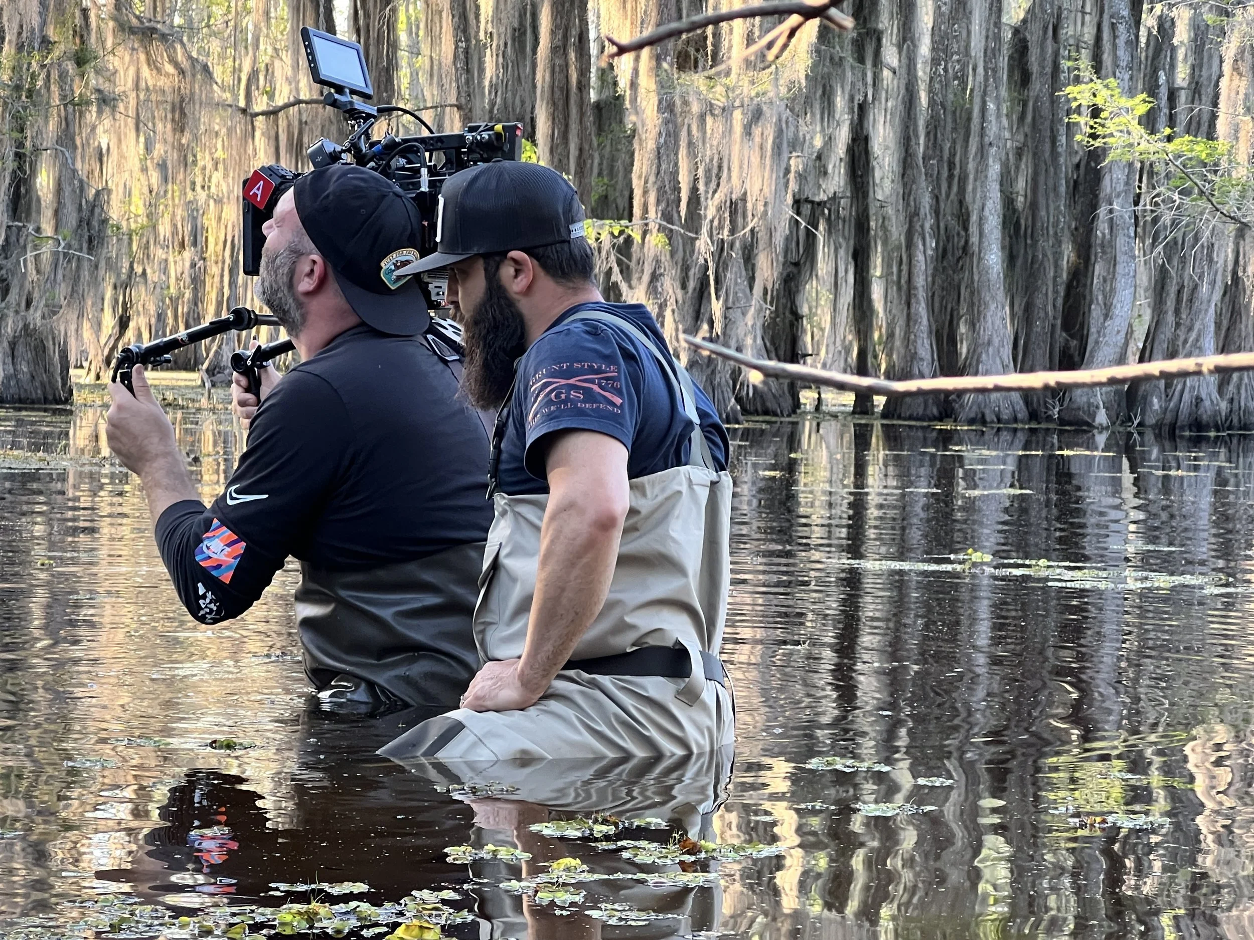 Two men are standing in the water of a swamp or marsh, operating a camera mounted on a stabilizer. They are surrounded by tall trees with Spanish moss hanging from the branches, and the water is calm with some floating leaves and small plants.