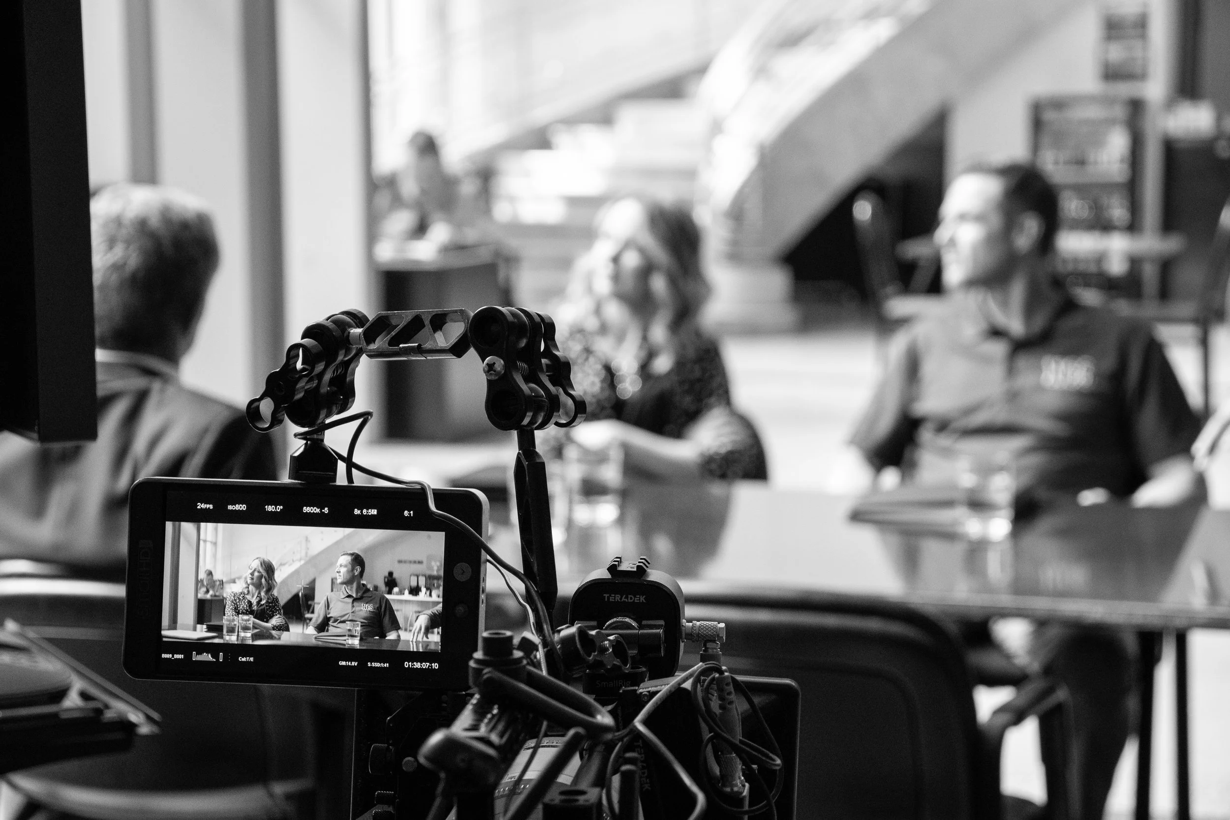 On a film set, a camera monitors two people in a discussion, with a woman and a man seated at a table, in front of a blurred background.