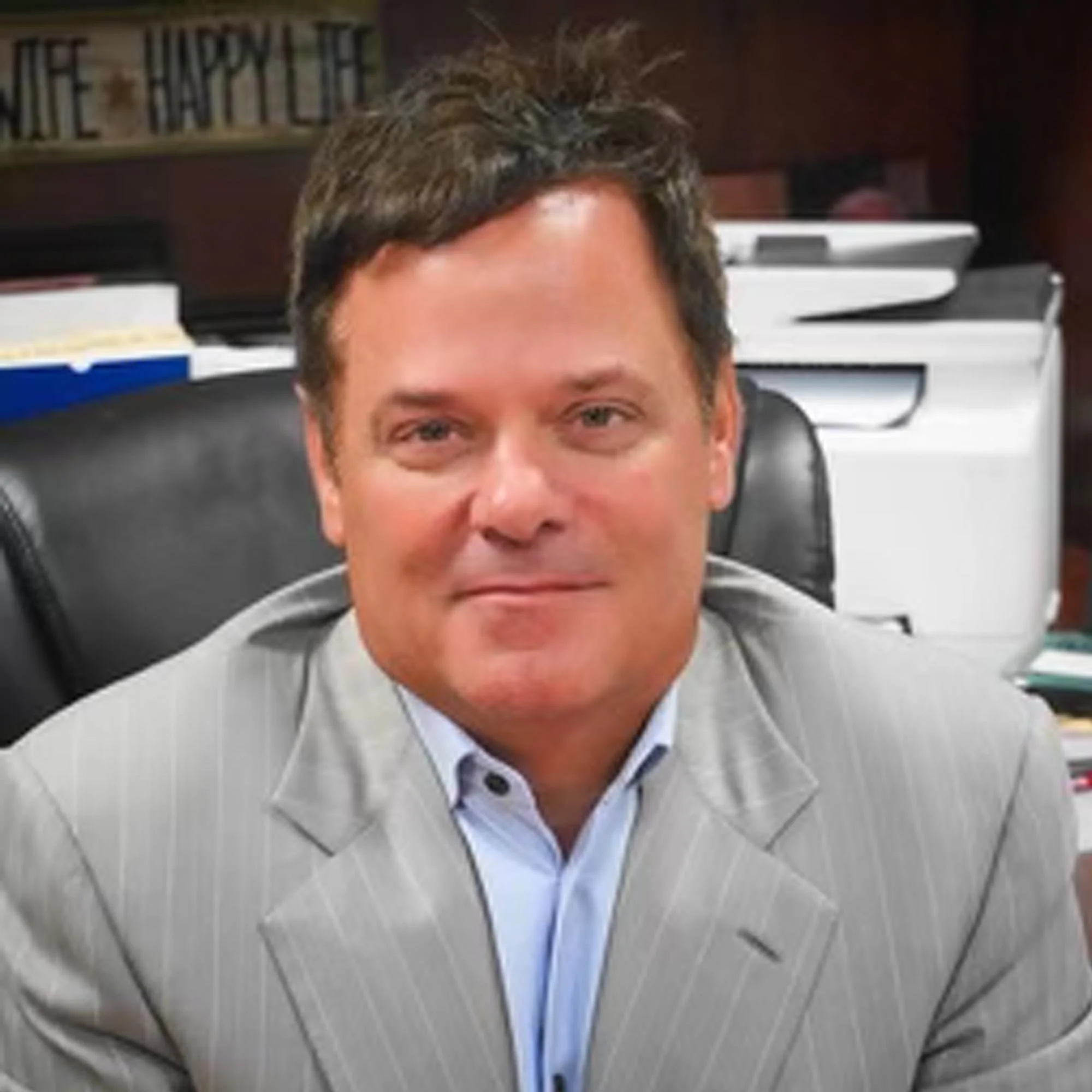 A man sitting at a desk in an office, wearing a light gray pinstripe suit and a white shirt, with books and papers behind him.