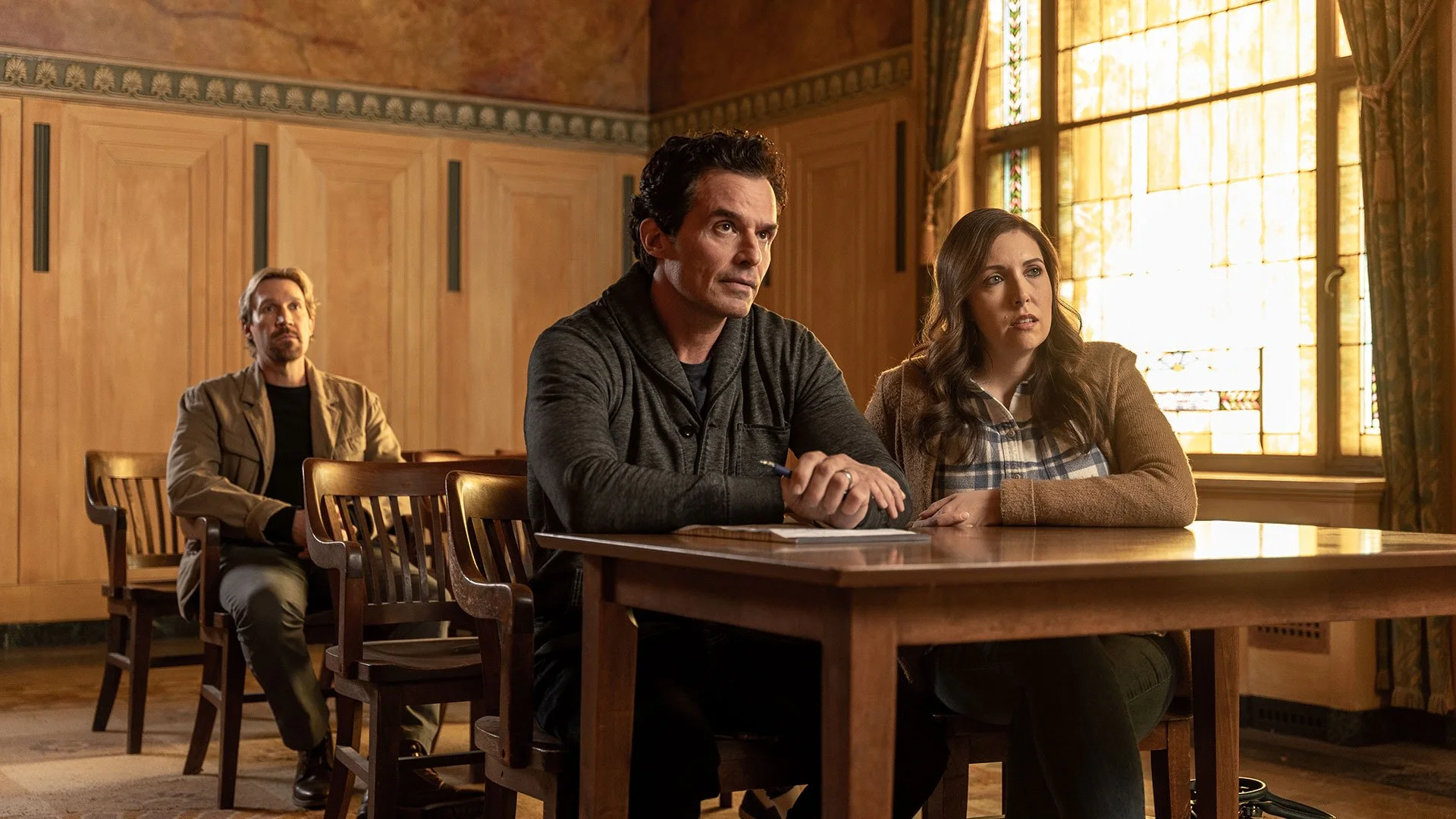 Three people sitting at a wooden table in a room with large windows and stained glass, engaged in a serious conversation.