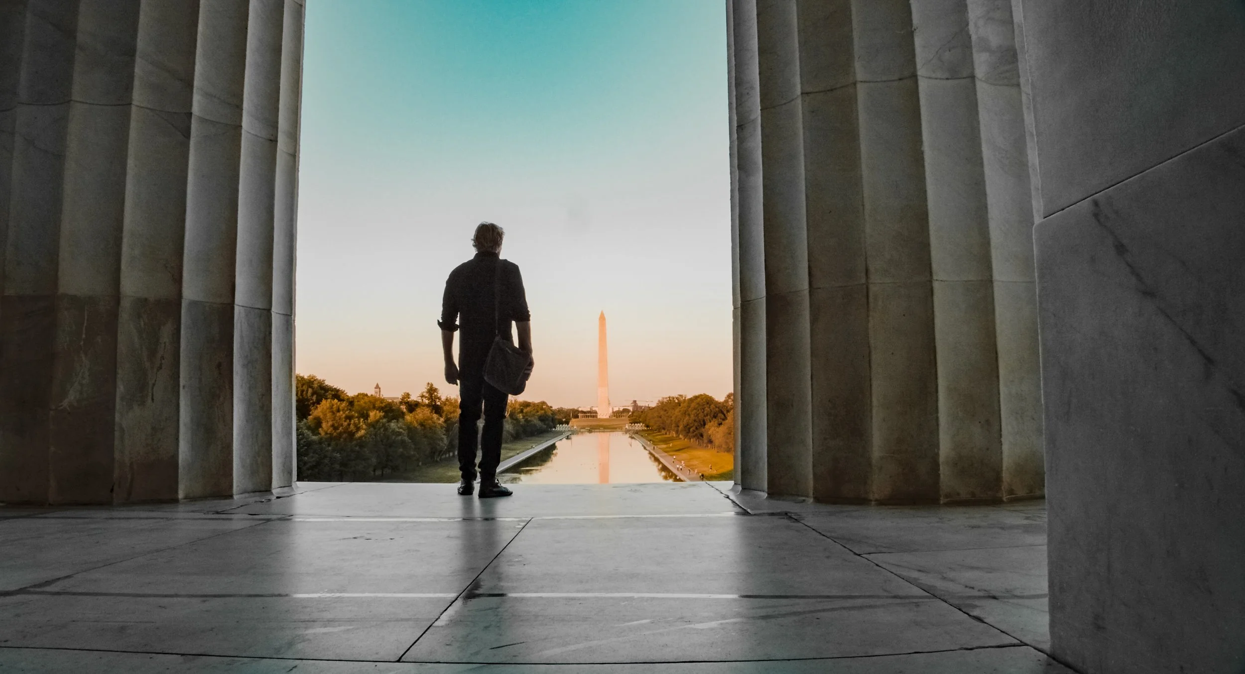 Silhouette of a man standing at the Lincoln Memorial, looking out at the Washington Monument and reflecting pool at sunset.