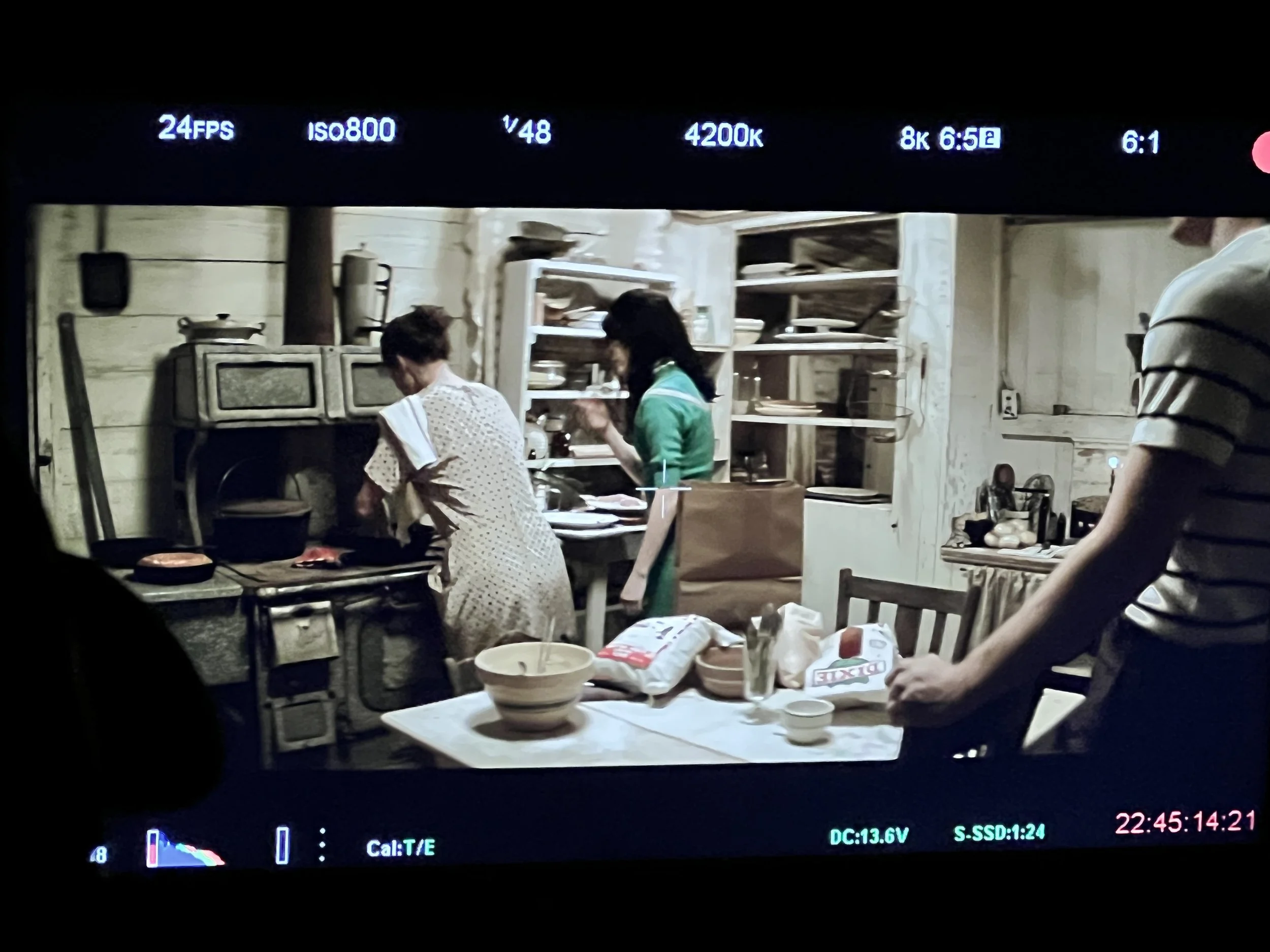 Two women cooking in a rustic kitchen, one in a white polka dot dress and one in a green outfit, with a table of bowls and bags of ingredients in the foreground.