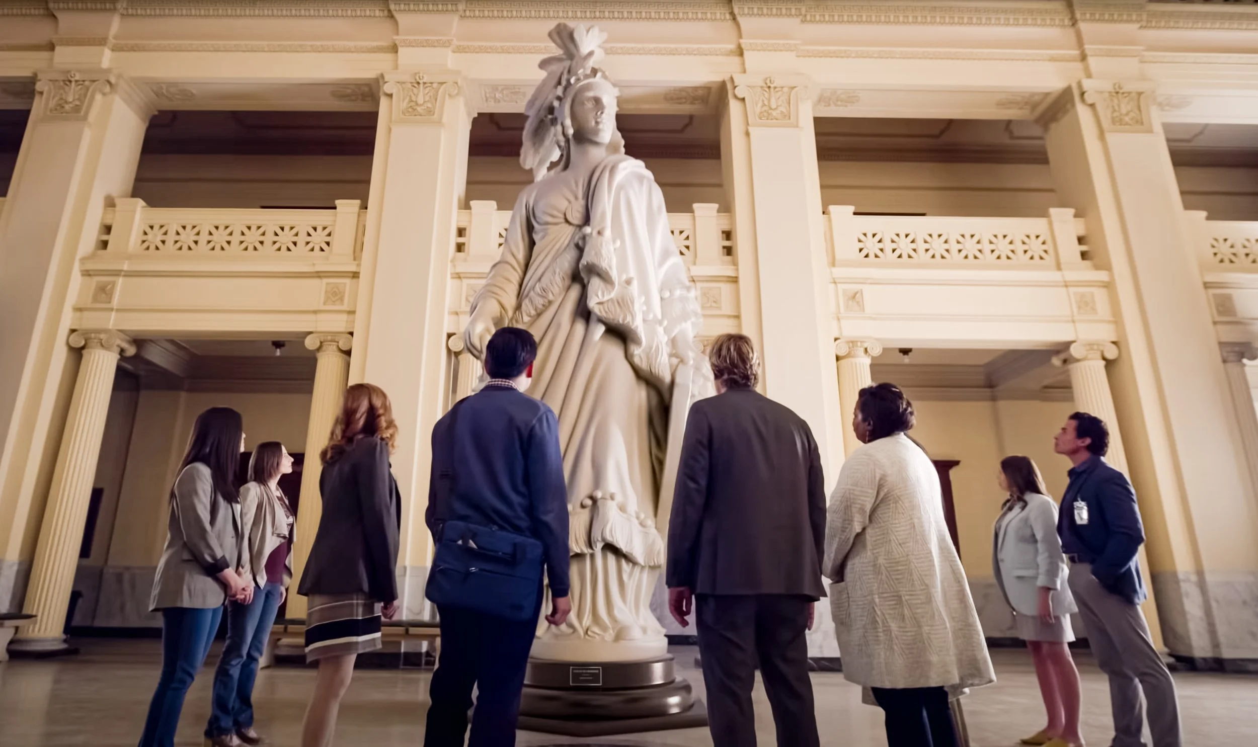 Group of people viewing a large classical statue of a historic figure in an elegant museum hall with columns and ornate railings.