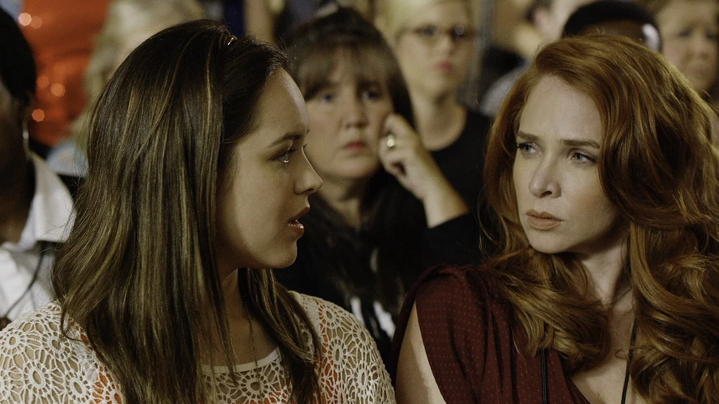 Close-up of two women with serious expressions sitting in an audience, surrounded by other people.