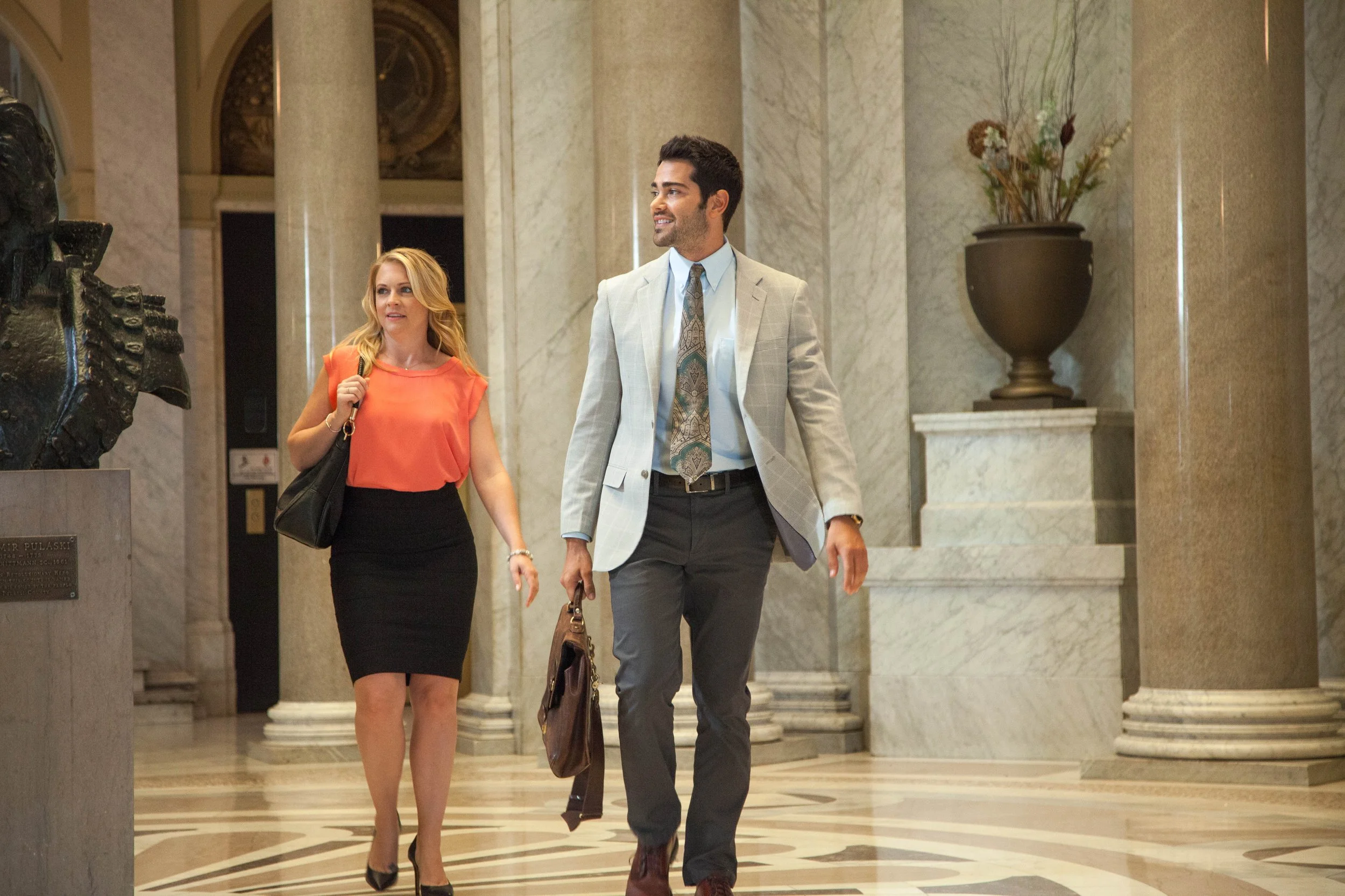 Two professionally dressed individuals, a woman and a man, walking through a luxurious marble building lobby with tall columns and artwork.