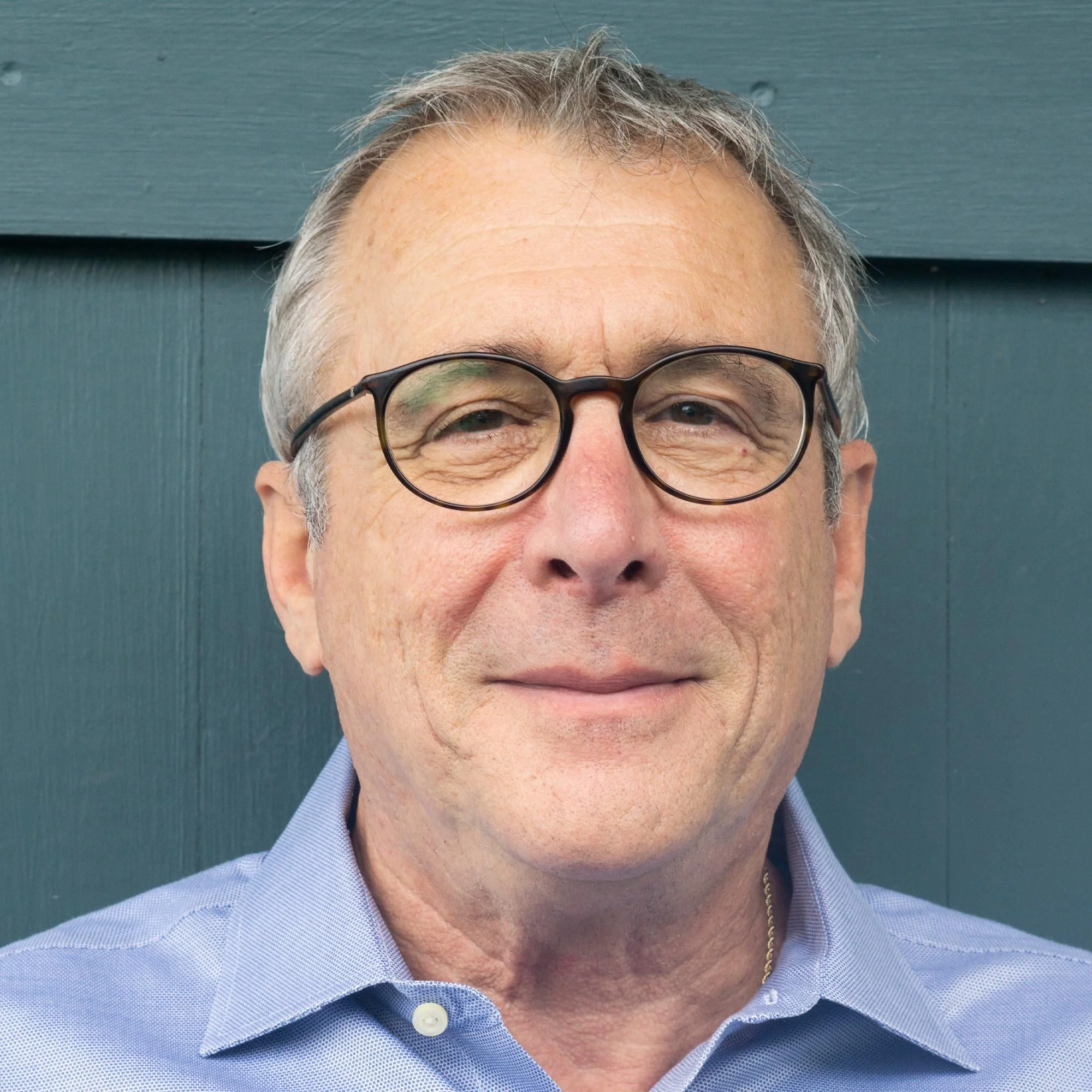 Close-up of a middle-aged man with gray hair wearing glasses and a light blue collared shirt, smiling in front of a blue-green wooden wall.
