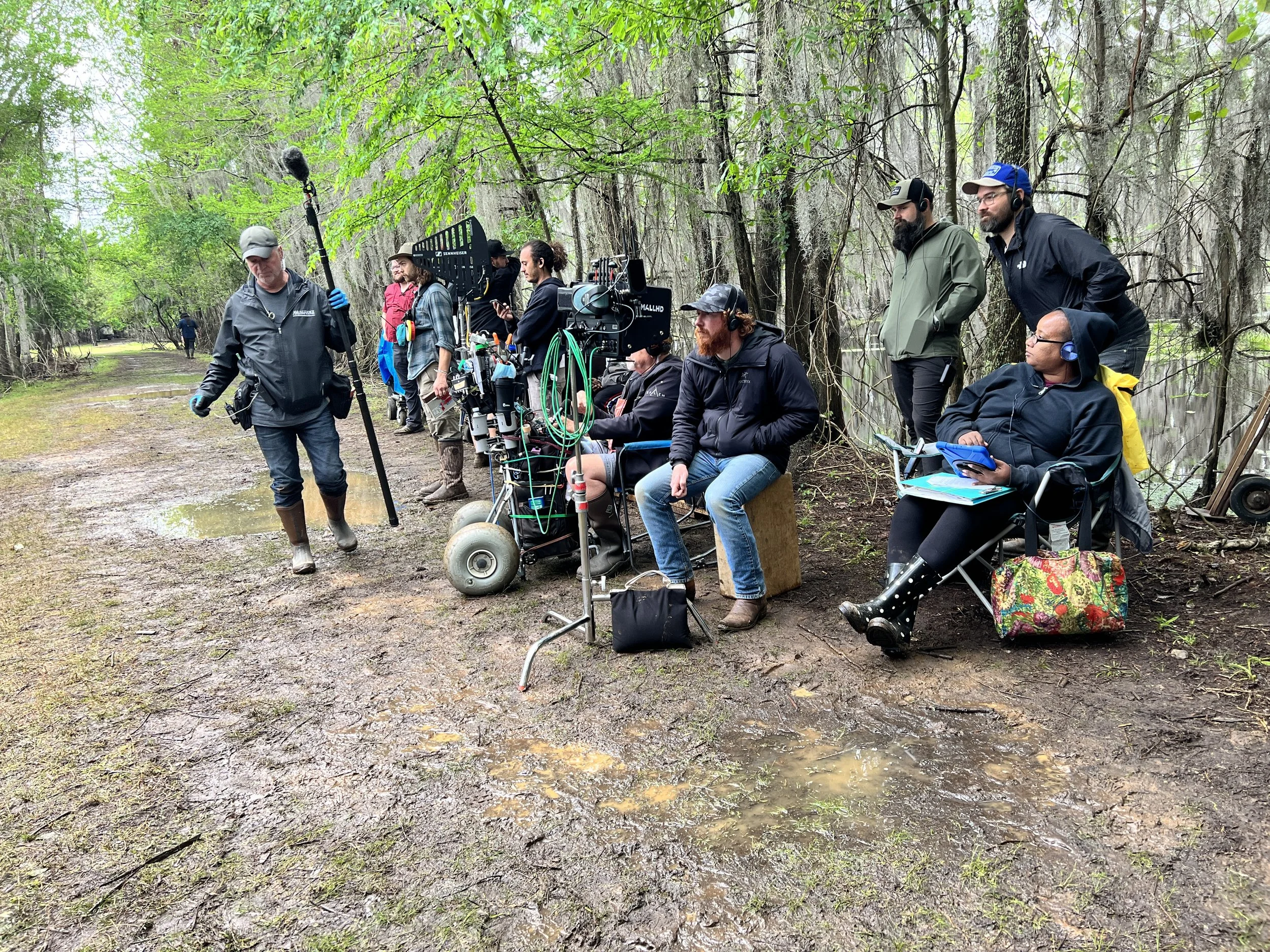 Film crew shooting in a forest, with a person holding a boom microphone, several crew members sitting and standing near camera equipment, and trees surrounding the scene.
