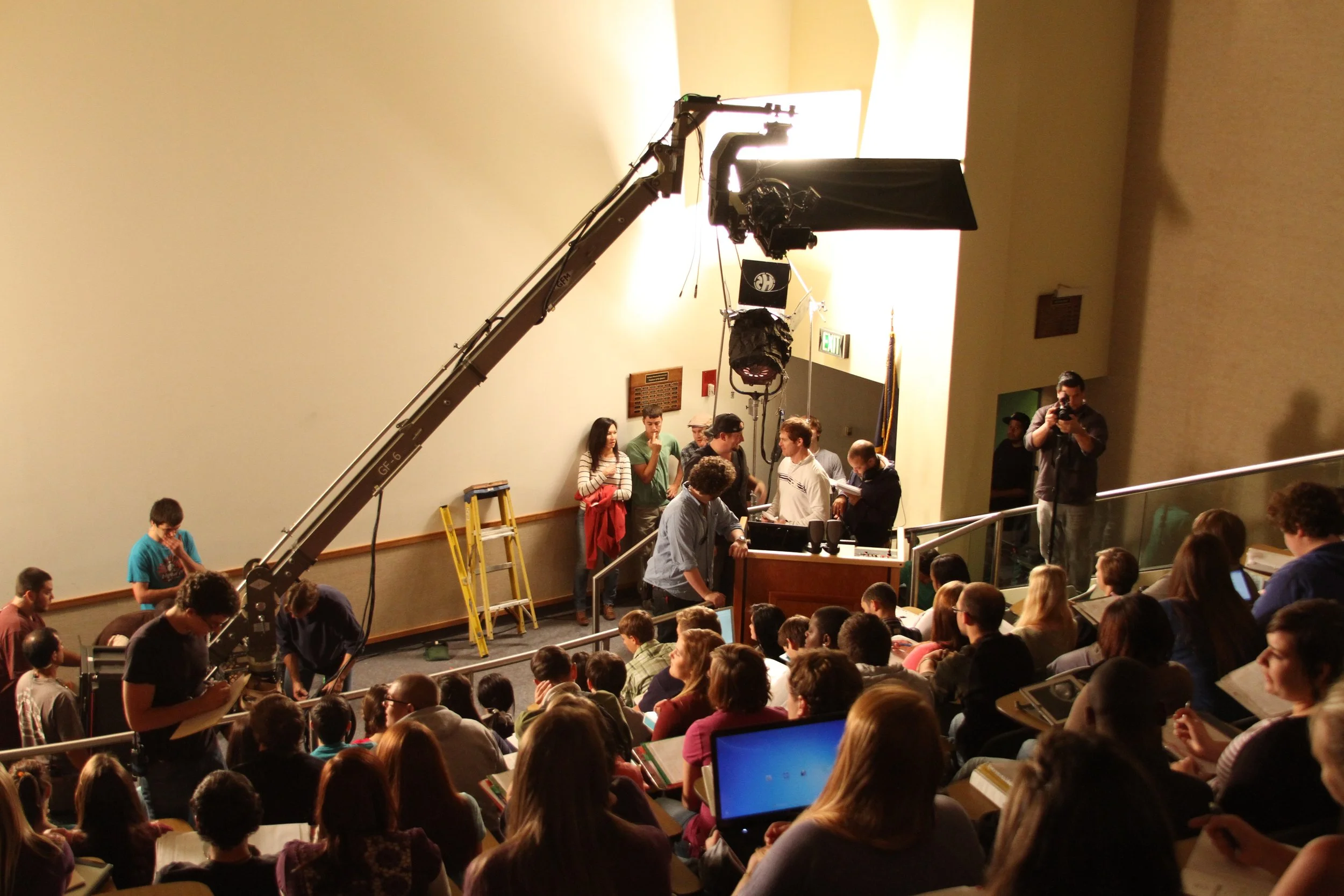 A large group of people gathered in an auditorium for a discussion or event. Crew members are operating camera equipment with a large crane and lighting. Some individuals are at a podium, while others are standing or seated, engaging with the event.