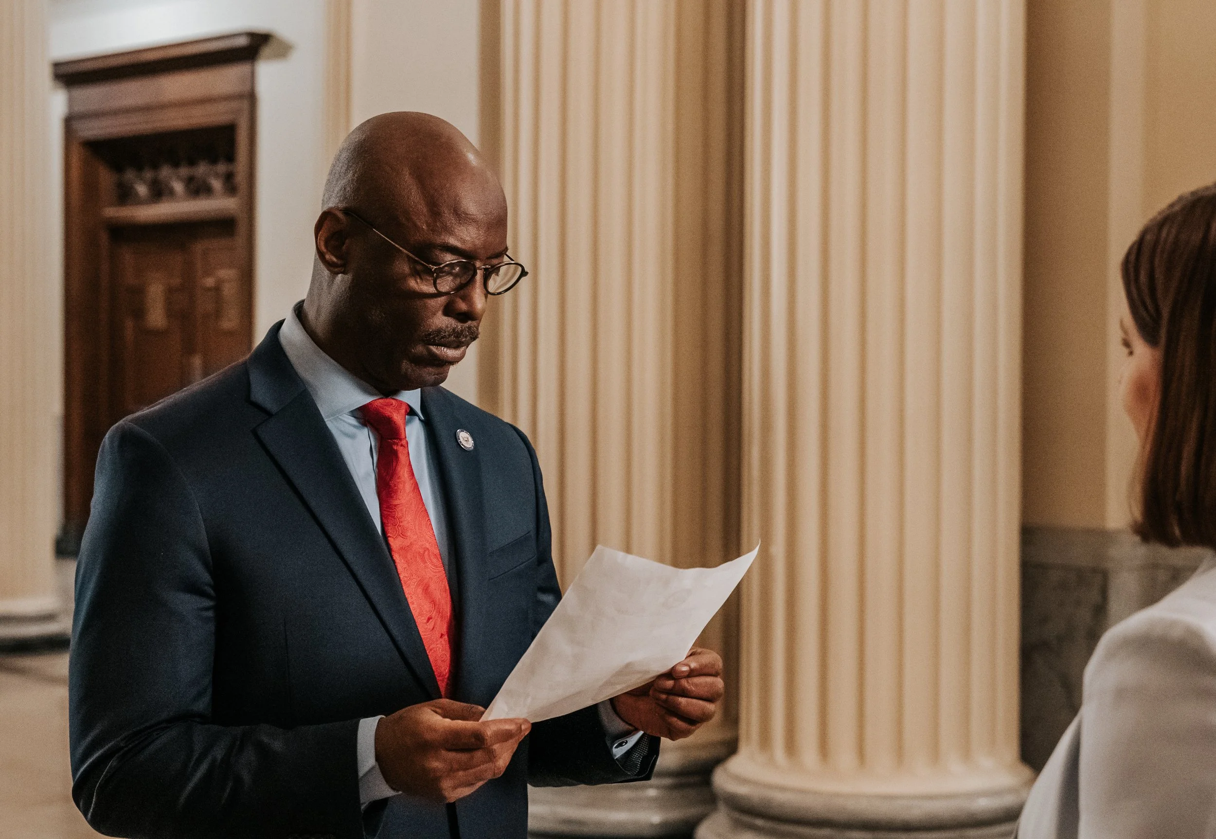 An older African American man in a dark suit, glasses, and a red tie, looking at a piece of paper while standing indoors near ornate columns.