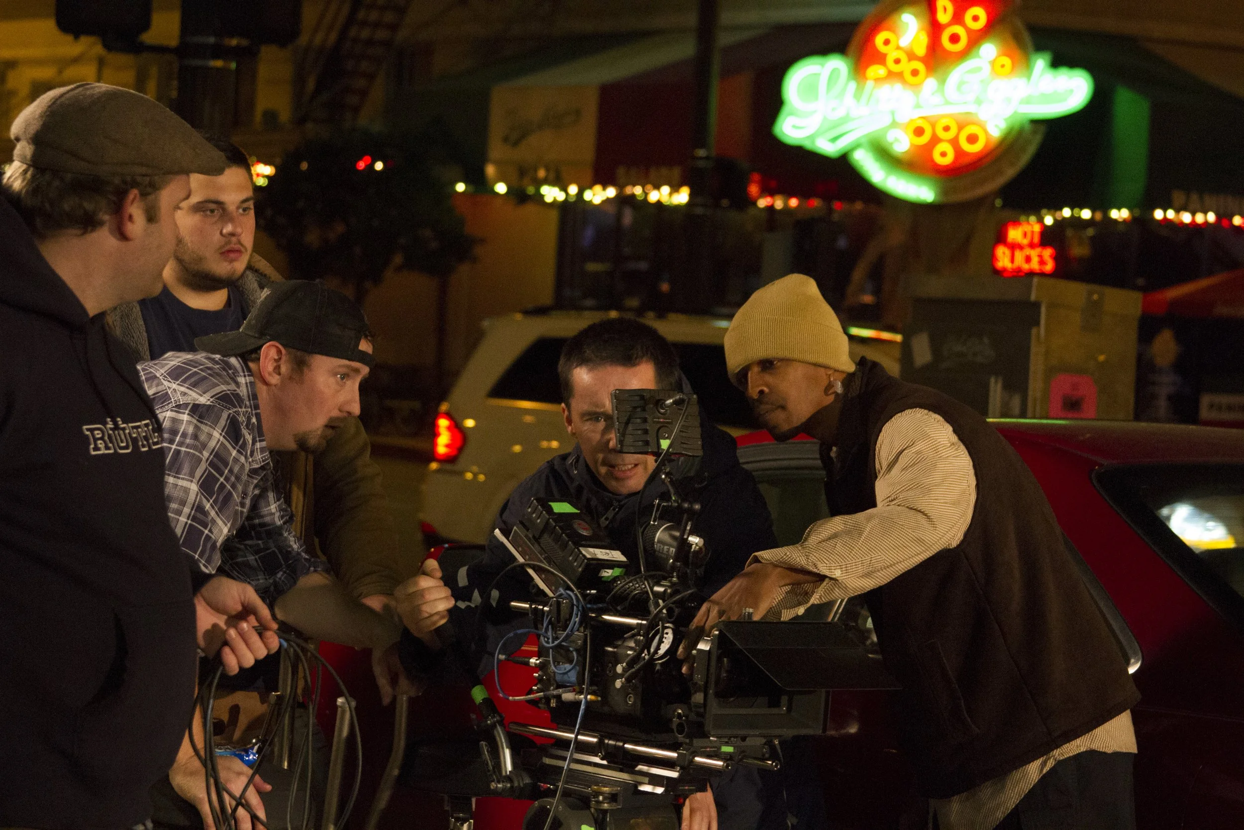 Group of people working with filming equipment outdoors at night, with neon signs and parked cars in the background.