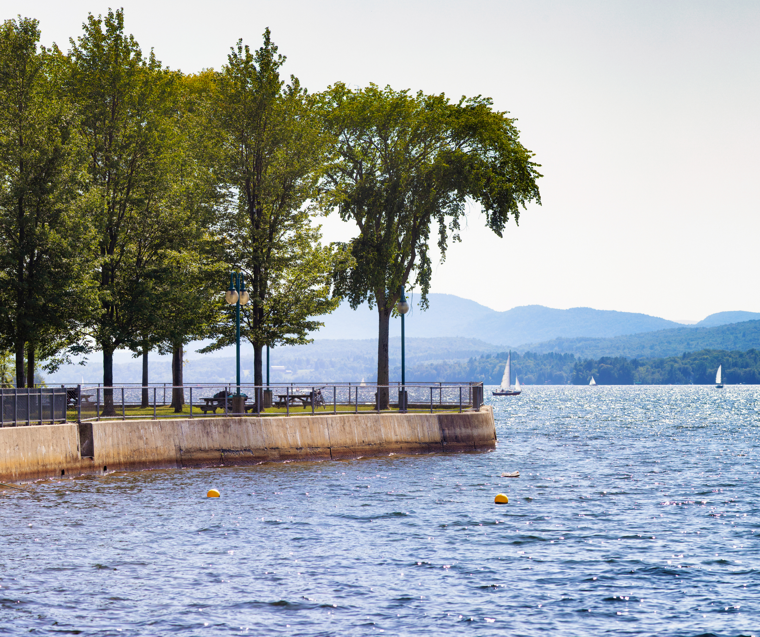Parc  Pointe-Merry à Magog. Quai au bord de l'eau avec des arbres, lampadaires, bancs, et voiliers au large, montagnes en arrière-plan.