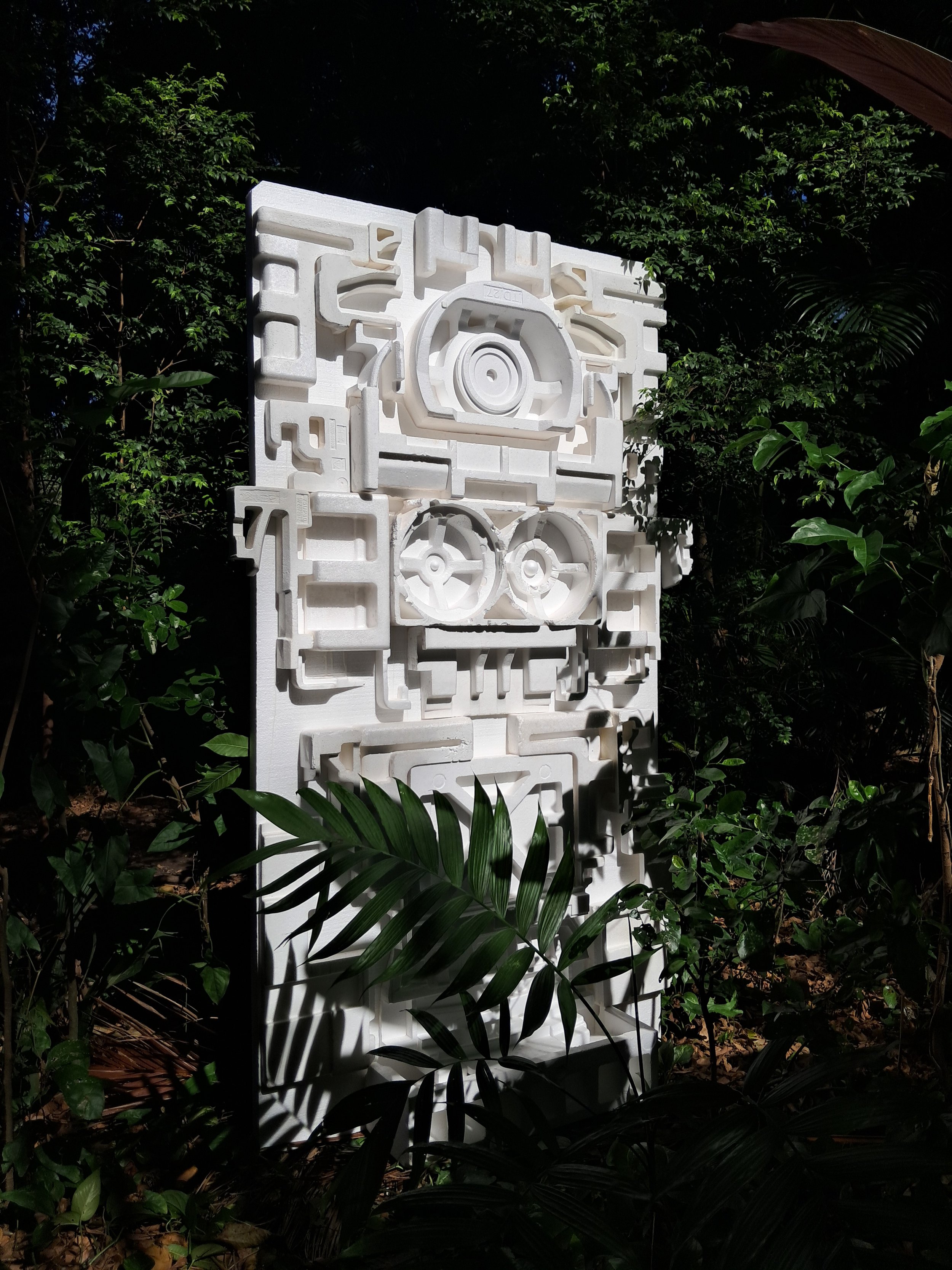 Escultura moderna blanca en forma de bloques y engranajes en un entorno rodeado de vegetación verde.