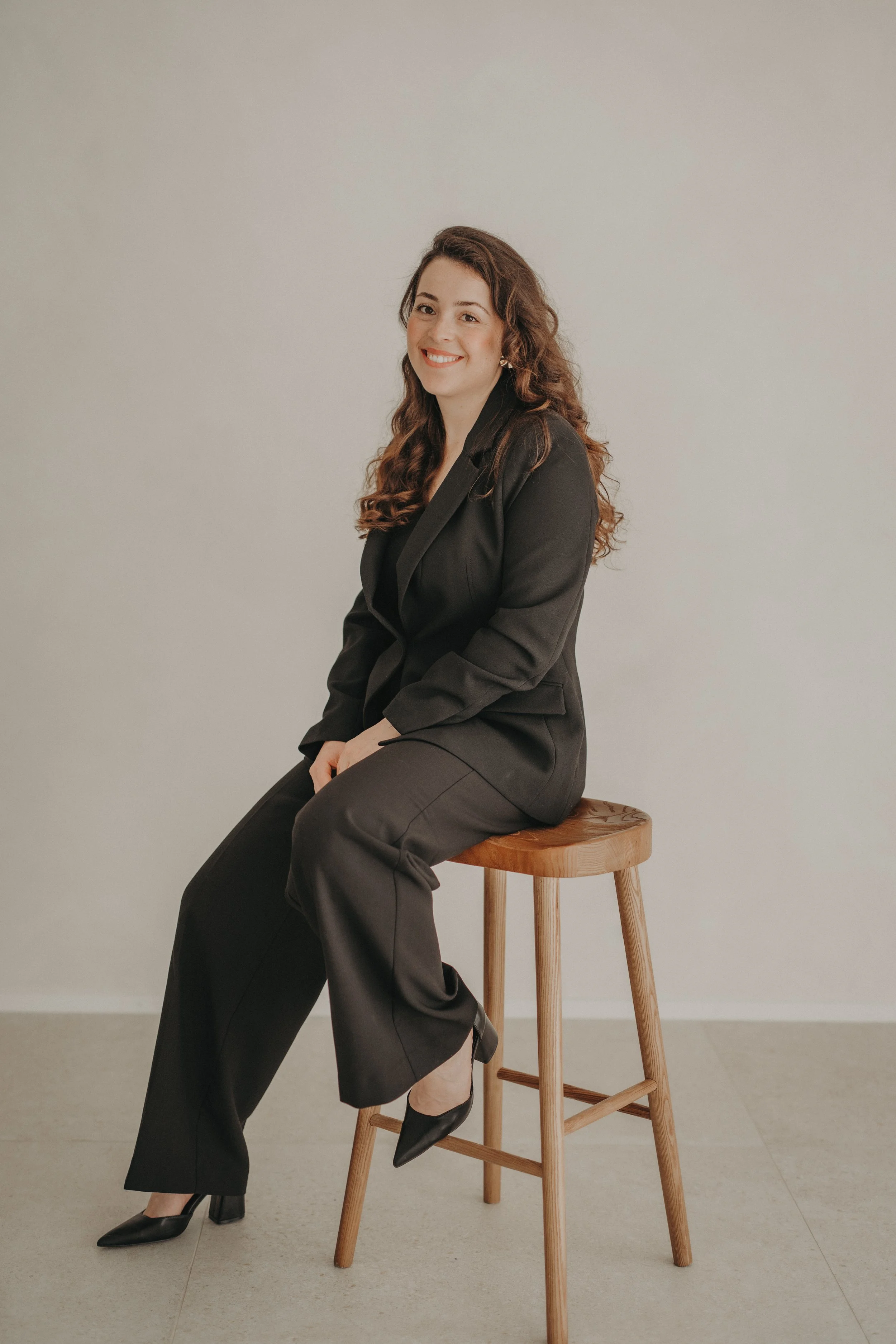 Une femme assise sur un tabouret en bois, portant un tailleur noir, avec des chaussures à talons noirs, souriant et regardant vers l'objectif, dans un décor minimaliste avec un mur blanc.