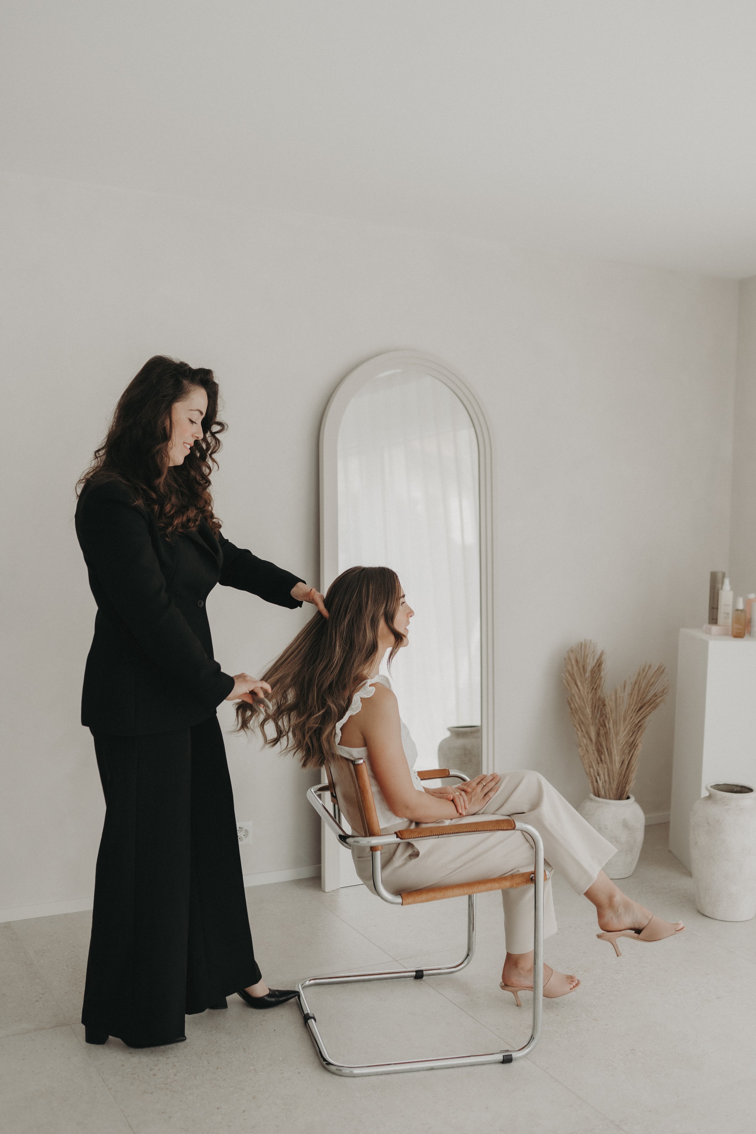Une femme assise sur une chaise en parlant avec une esthéticienne qui coiffe ses cheveux dans un salon de beauté minimaliste avec un miroir en arrière-plan.