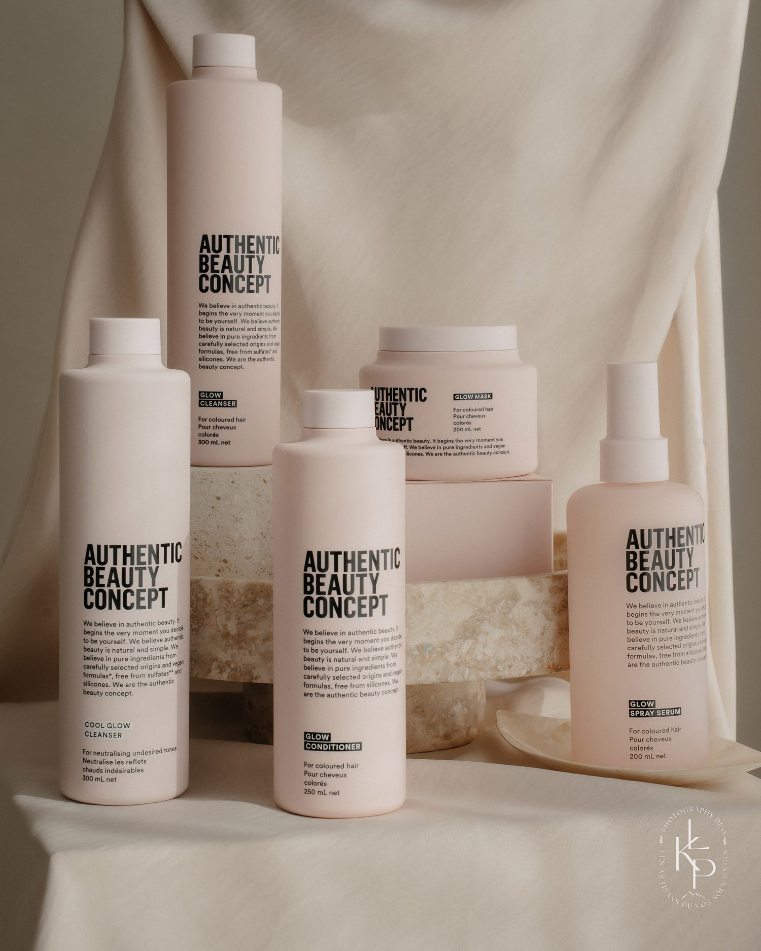 Gamme de produits de beauté 'Authentic Beauty Concept', comprenant un nettoyant, un conditionneur, un masque, un spray sérum, tous dans des contenants blancs avec étiquettes noires, disposés sur une surface en pierre beige, avec un fond de tissu blanc.